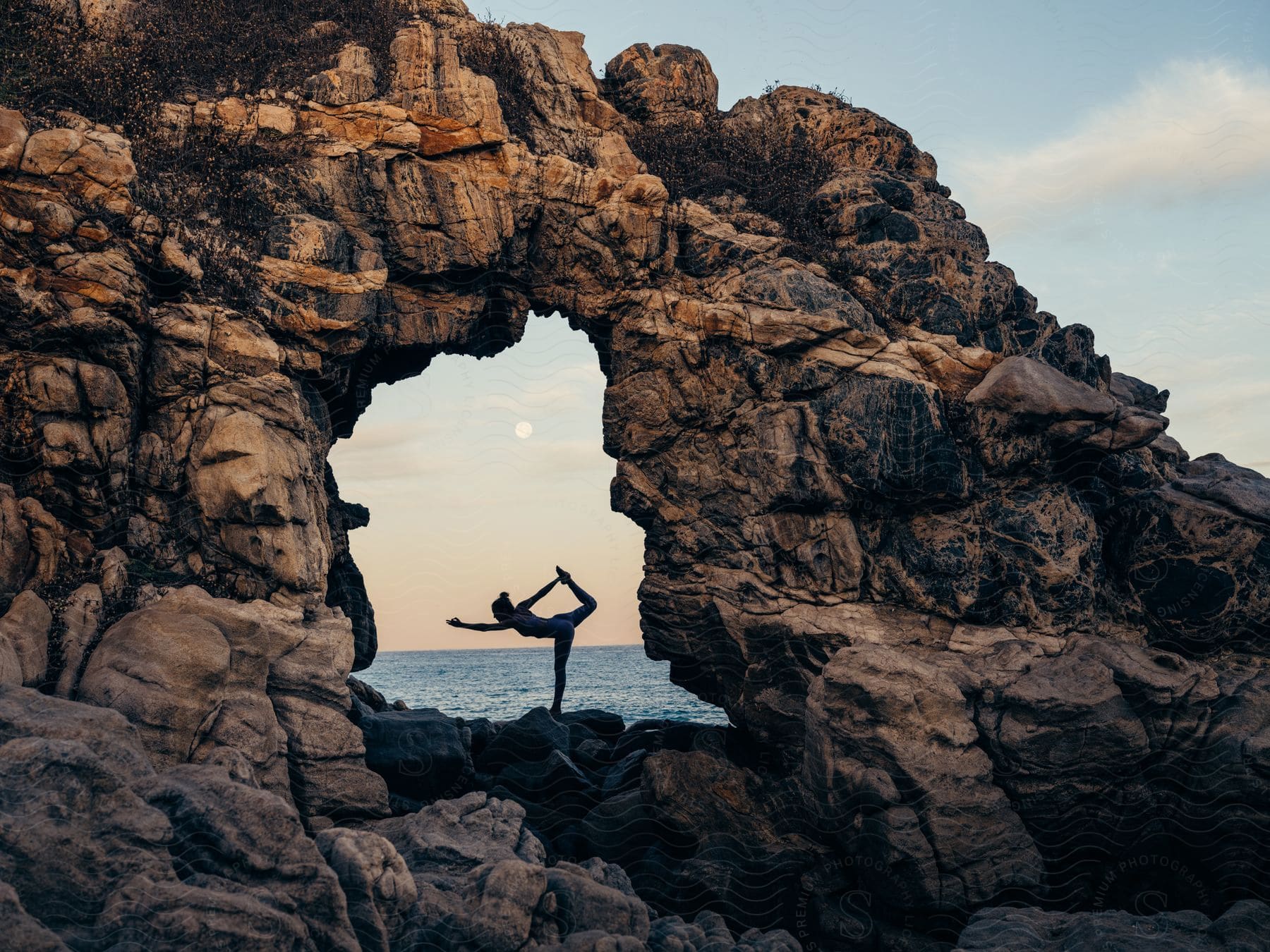 Woman balances in yoga pose on other side of opening in a coastal rock formation.