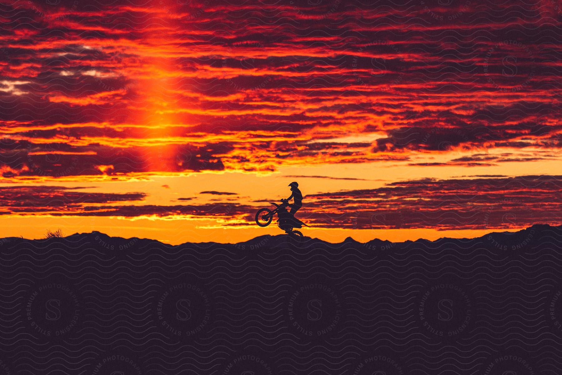 An off-road motorcyclist popping a wheeling in a hilly landscape.