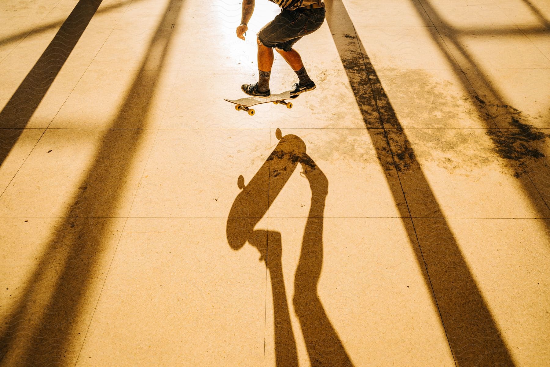 The feet of a man wearing shorts, socks, and a shoe are visible as he performs a skateboard stunt. The sun's rays reflect shadows of the building beams on the floor