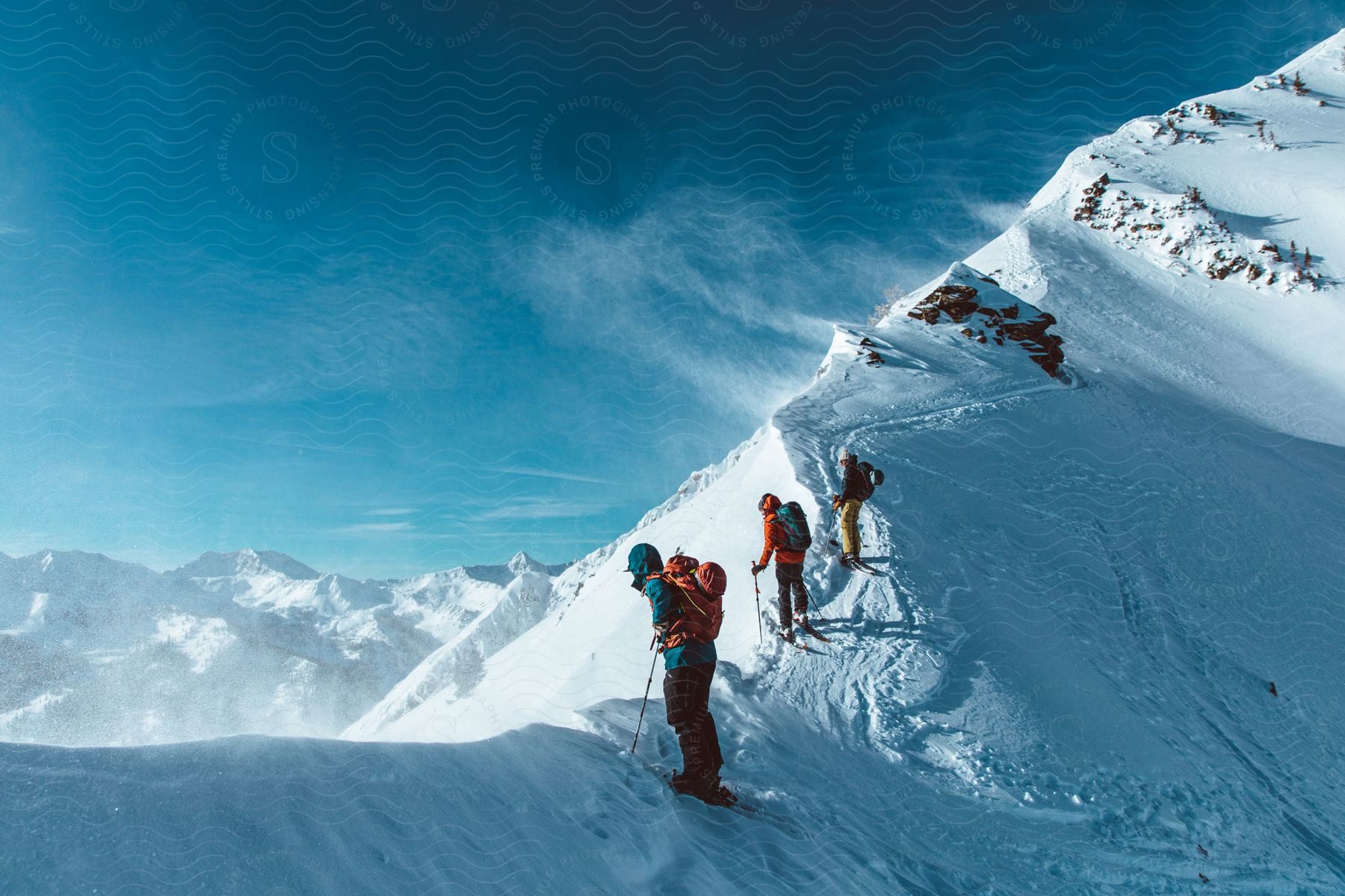 3 skiers with backpacks are at a top of a mountain.