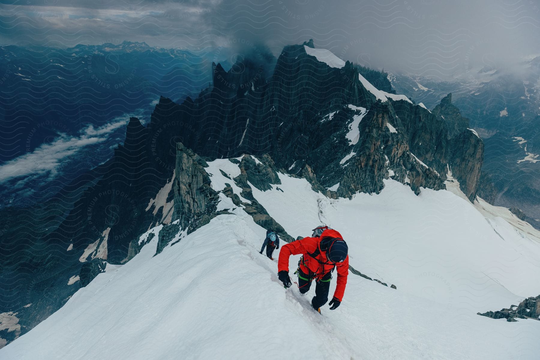 Two mountain climbers on a snow covered ridge by jagged rocky mountain peaks with a valley below under a dark clouded sky.