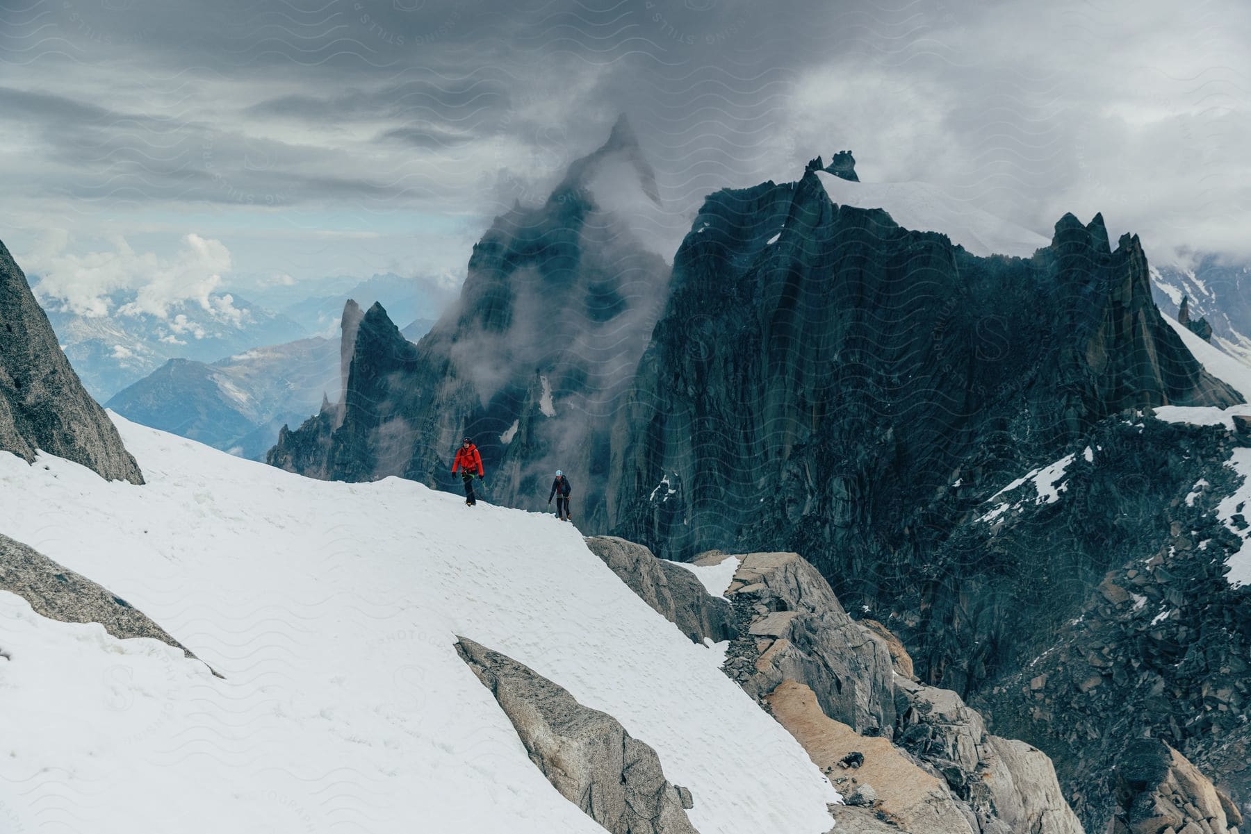 Two people are hiking at a high elevation on a mountain with snow on a cloudy day with a mountainous landscape in the background.