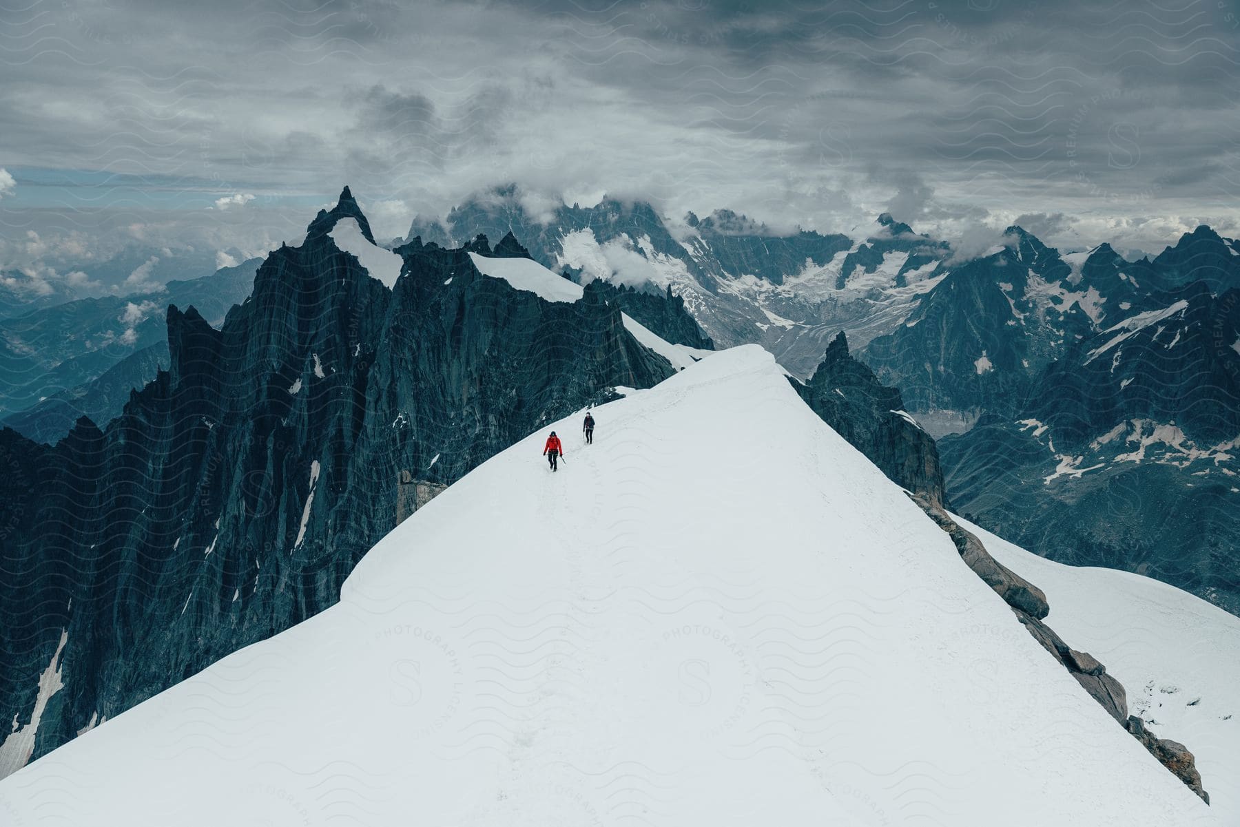 Two people are walking across a snow covered mountain near mountains with rough peaks high in a cloudy sky