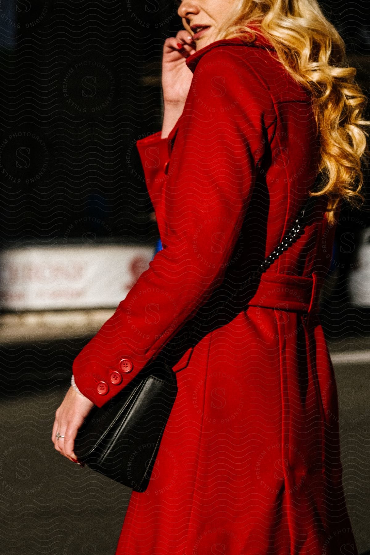 A blonde female wears a red coat and she has a black handbag throughout her shoulder.