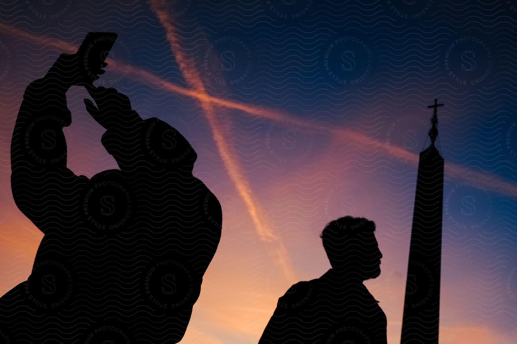 Silhouettes of two people standing outside as one person holds his phone up as pink contrails cross the sky