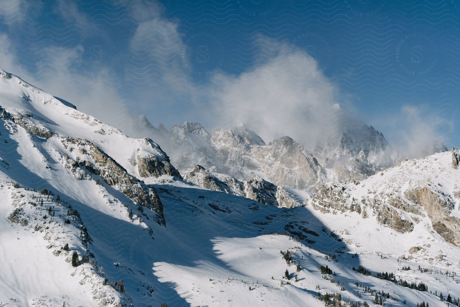 A snow-capped mountain, its peak shrouded in clouds, rises above a valley with a few trees clinging to its slopes.