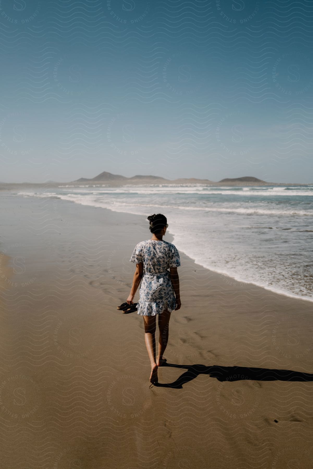 A woman walking along the waterline on a beach, sandals in hand and looking at the sea.