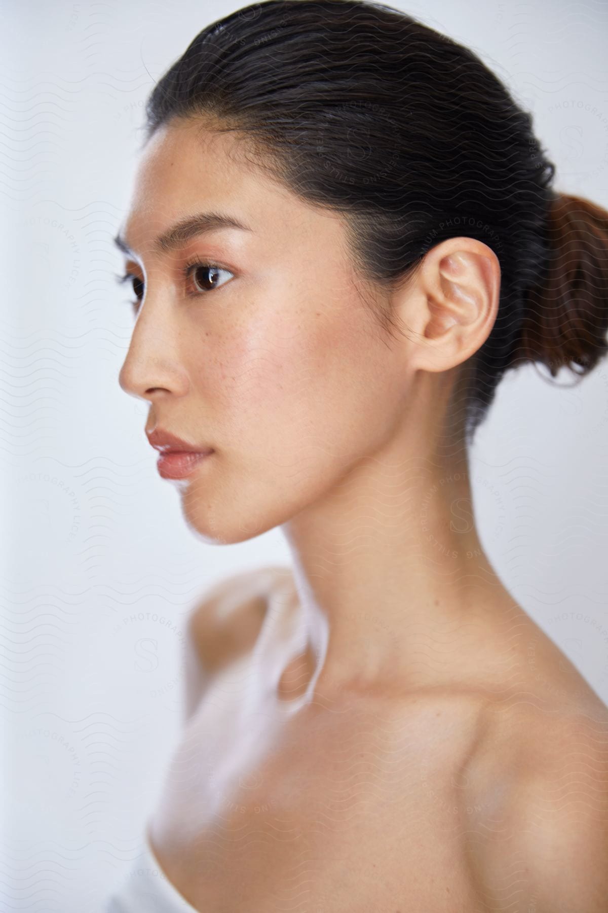 A Japanese woman in a white shoulderless top, her hair tied in a bun, looks off to the side with a serious expression.