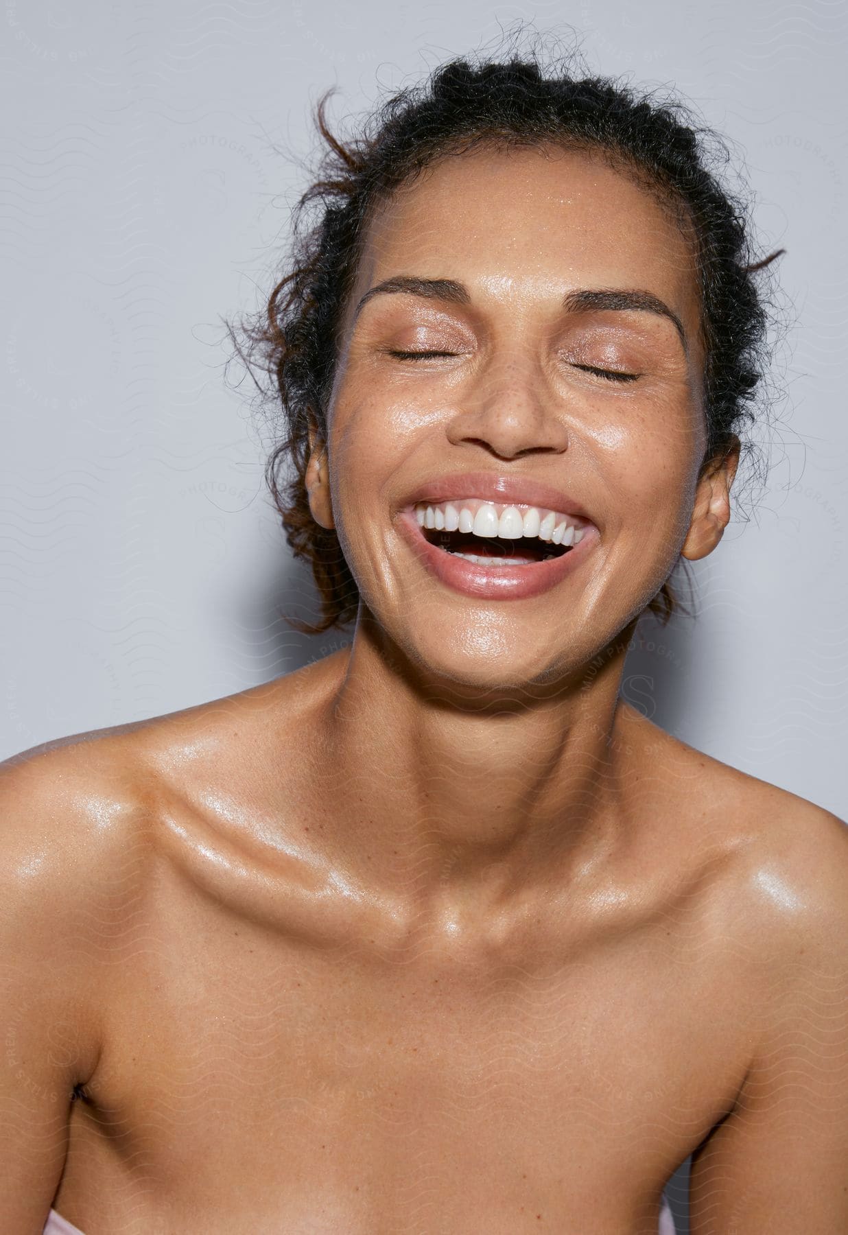 Woman with bare shoulders and damp skin laughs with her eyes closed.