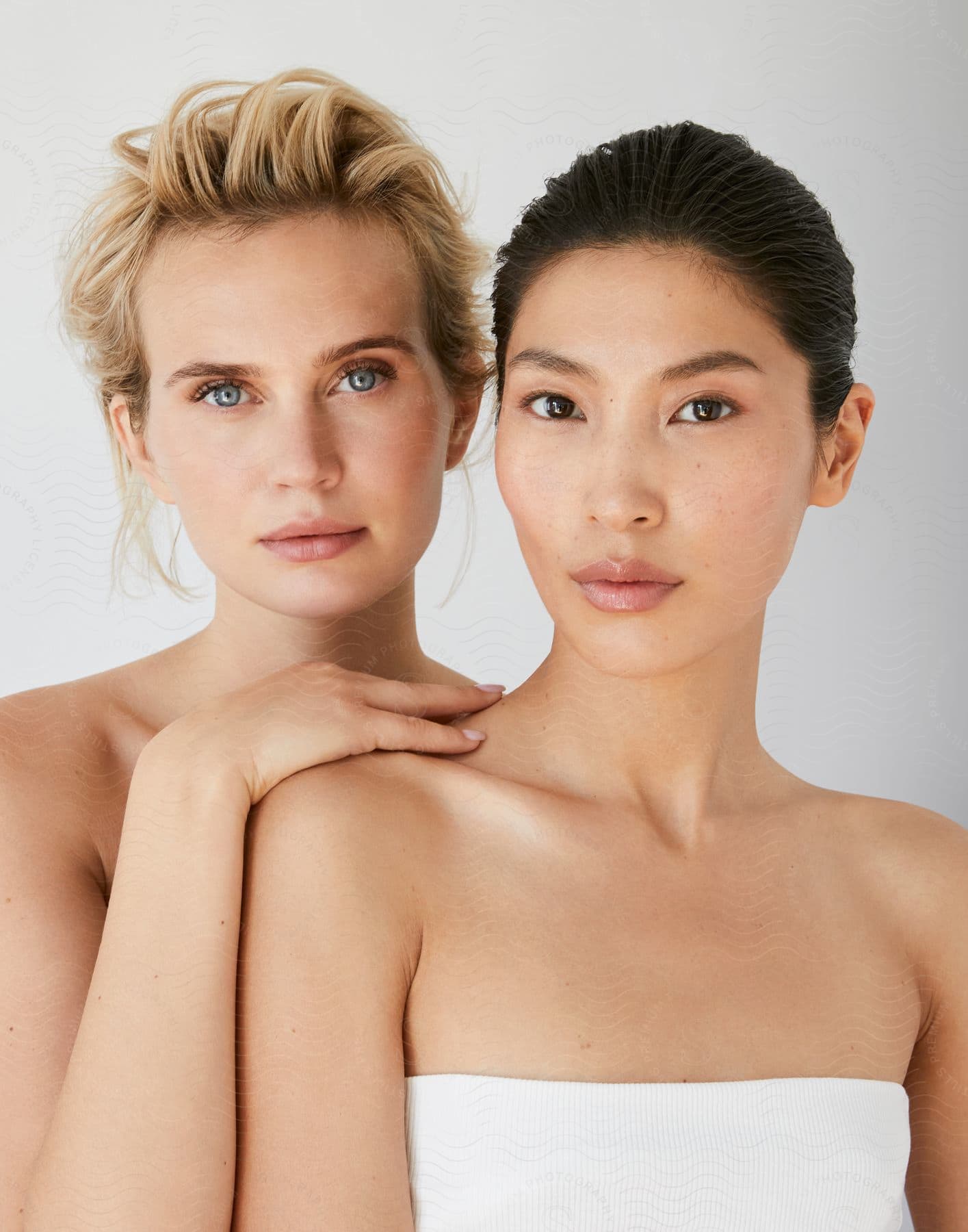 Two women of different ethnicities, one white with blond hair and the other Japanese with black hair, pose for a photo together, their bare shoulders exposed.