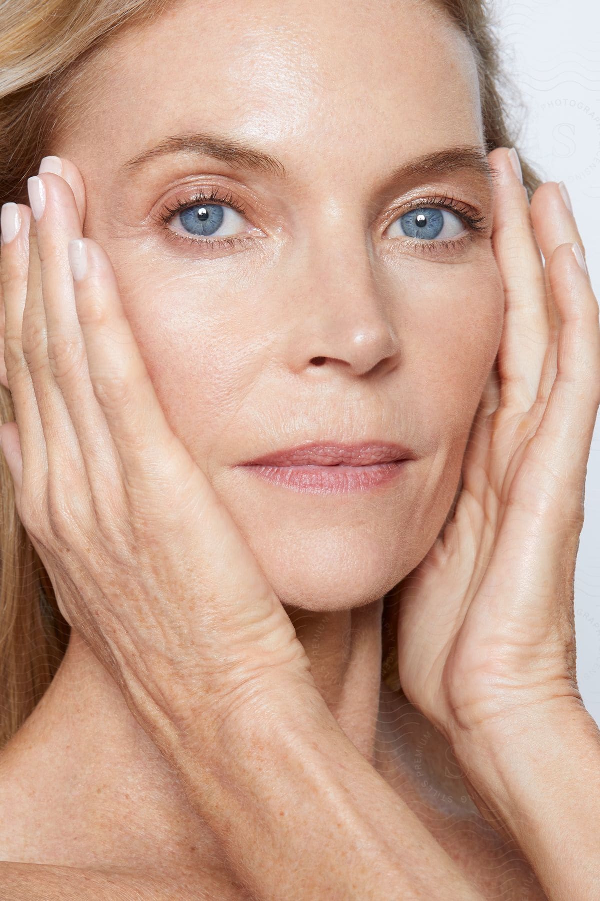 A close-up head shot of a mature woman with blond hair and blue eyes, her hands gently cupping her face.