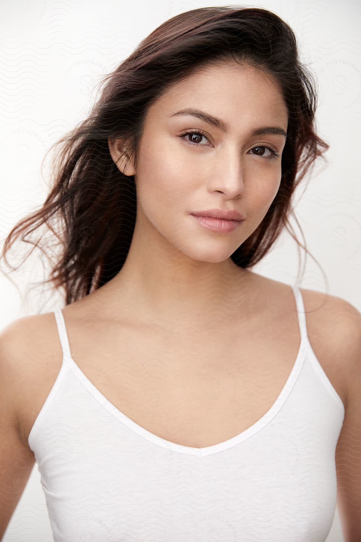 A Latina woman with brown hair and a white tank top stands against a white background