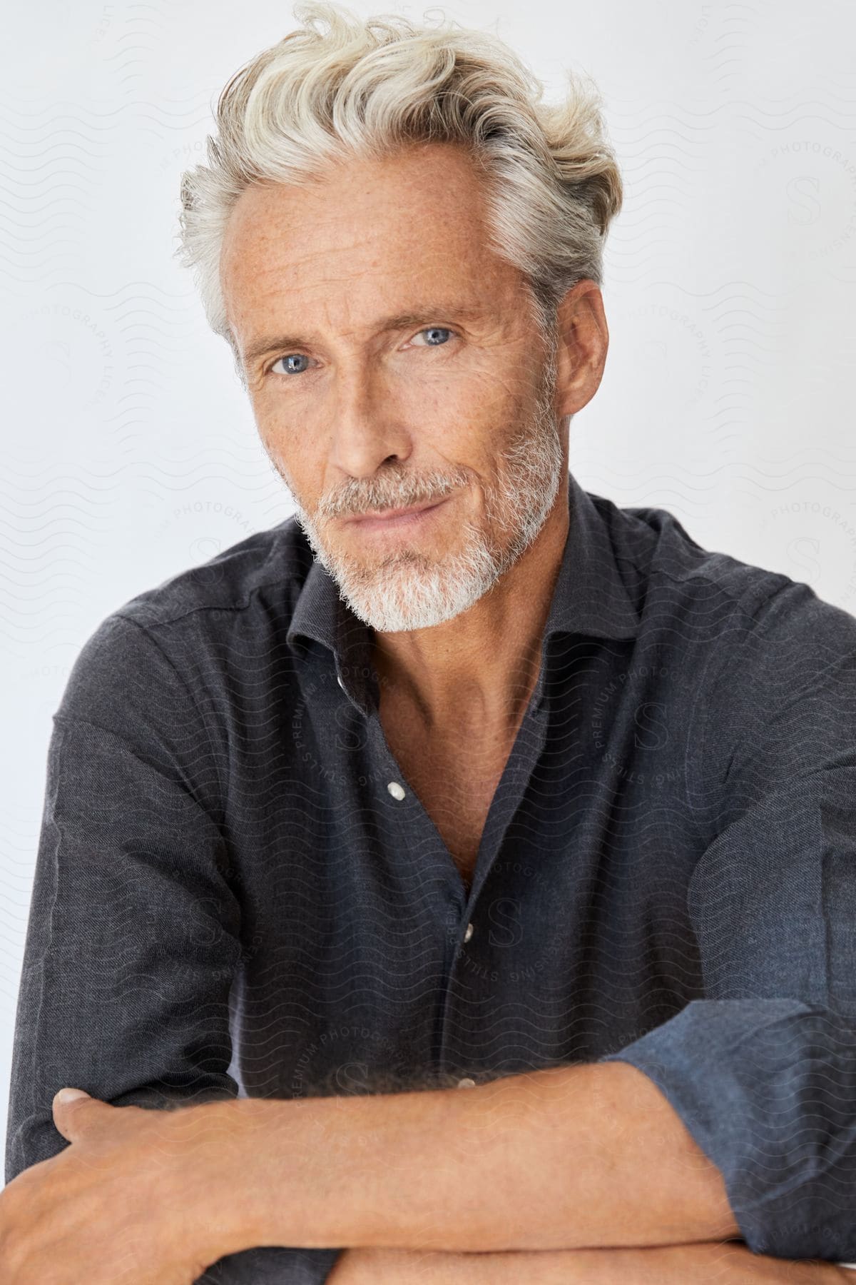 An older man with white and grey short wavy hair and a white goatee and short beard wearing a button up grey shirt