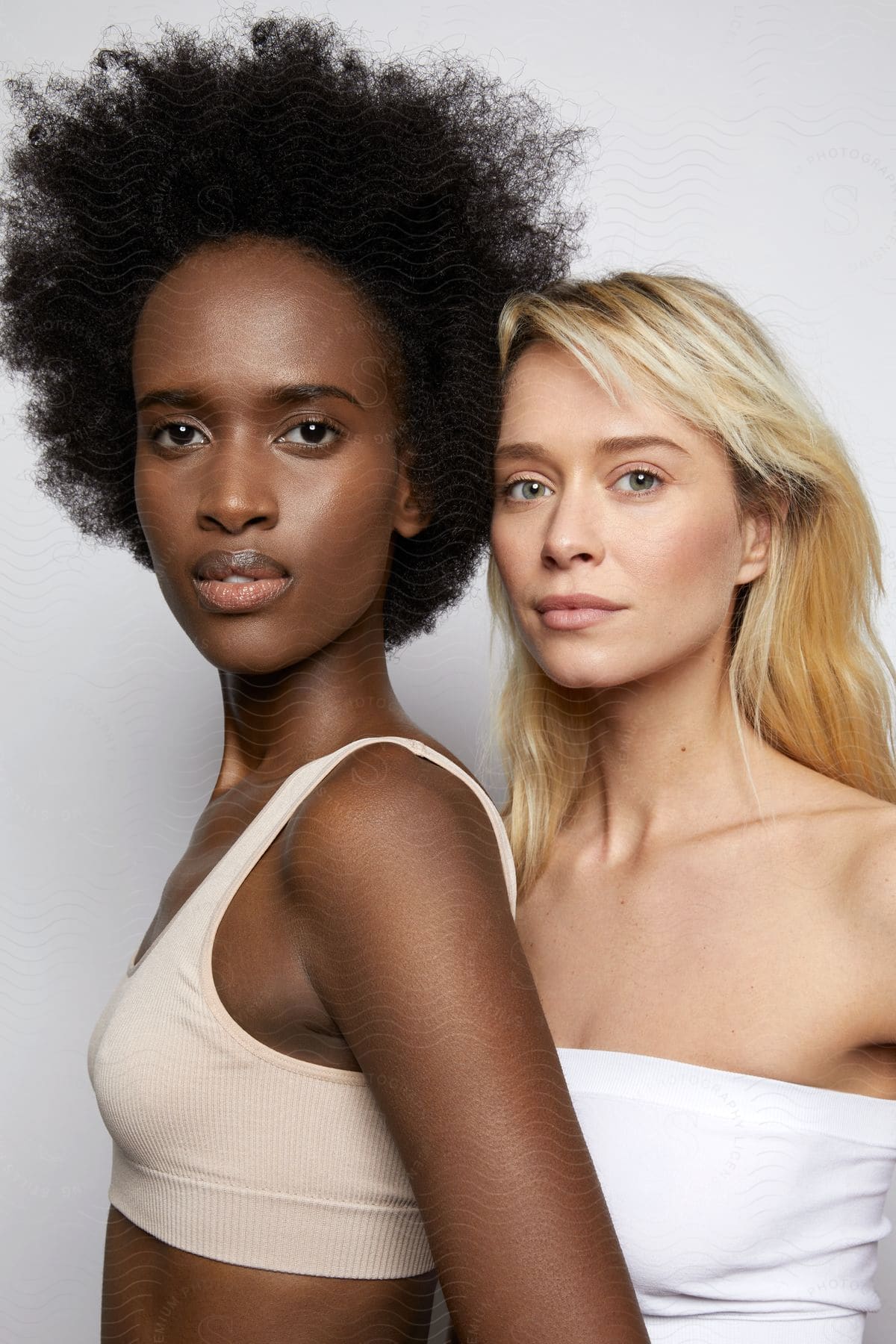 A white woman with blond hair and a black woman with black afro curly hair pose for a photo in one white shoulder less top and the other tan tank top.