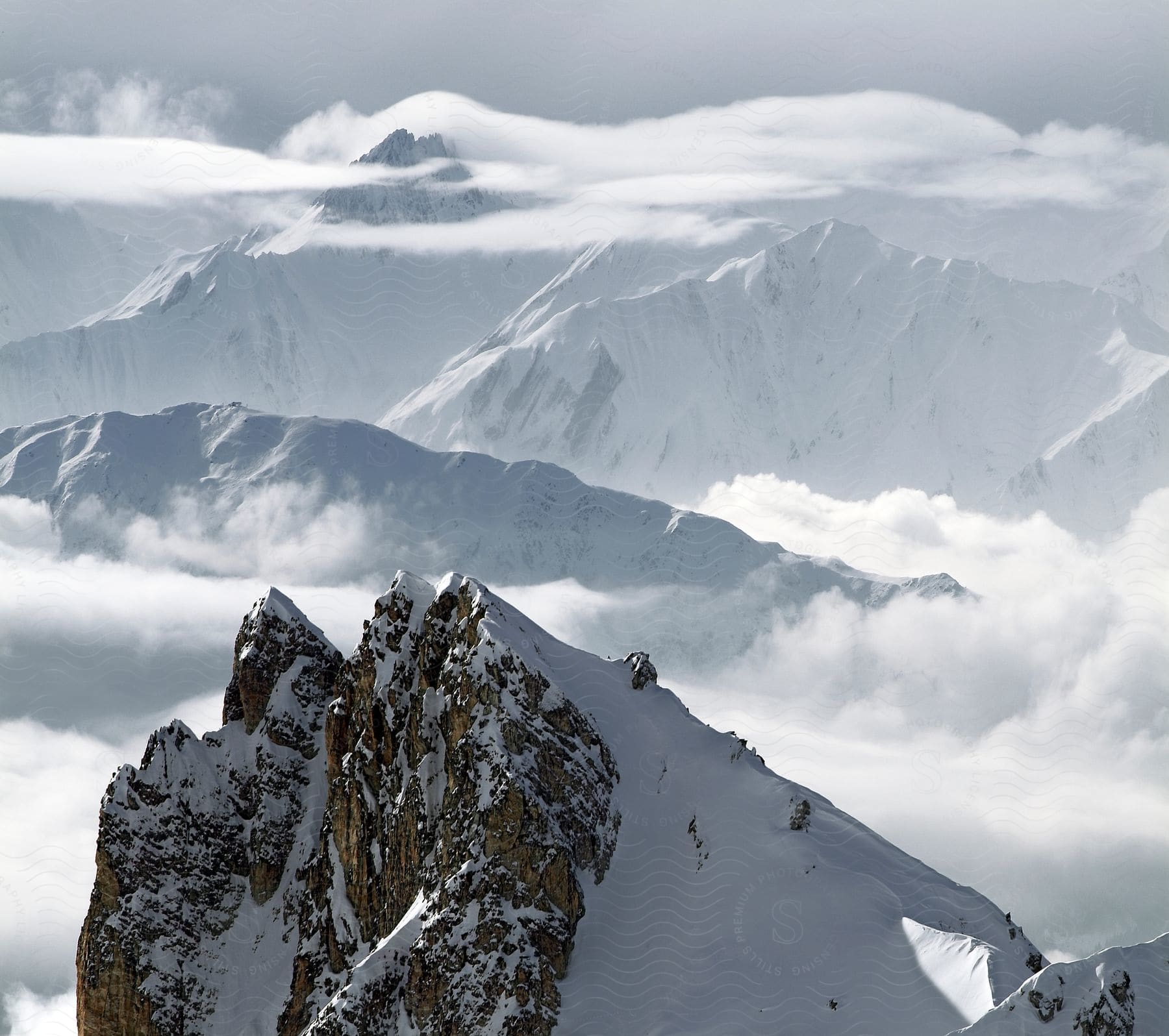 A snow-capped mountain range covered in clouds in the horizon, with jagged peaks and a foreground peak covered in snow, is seen from an aerial view.