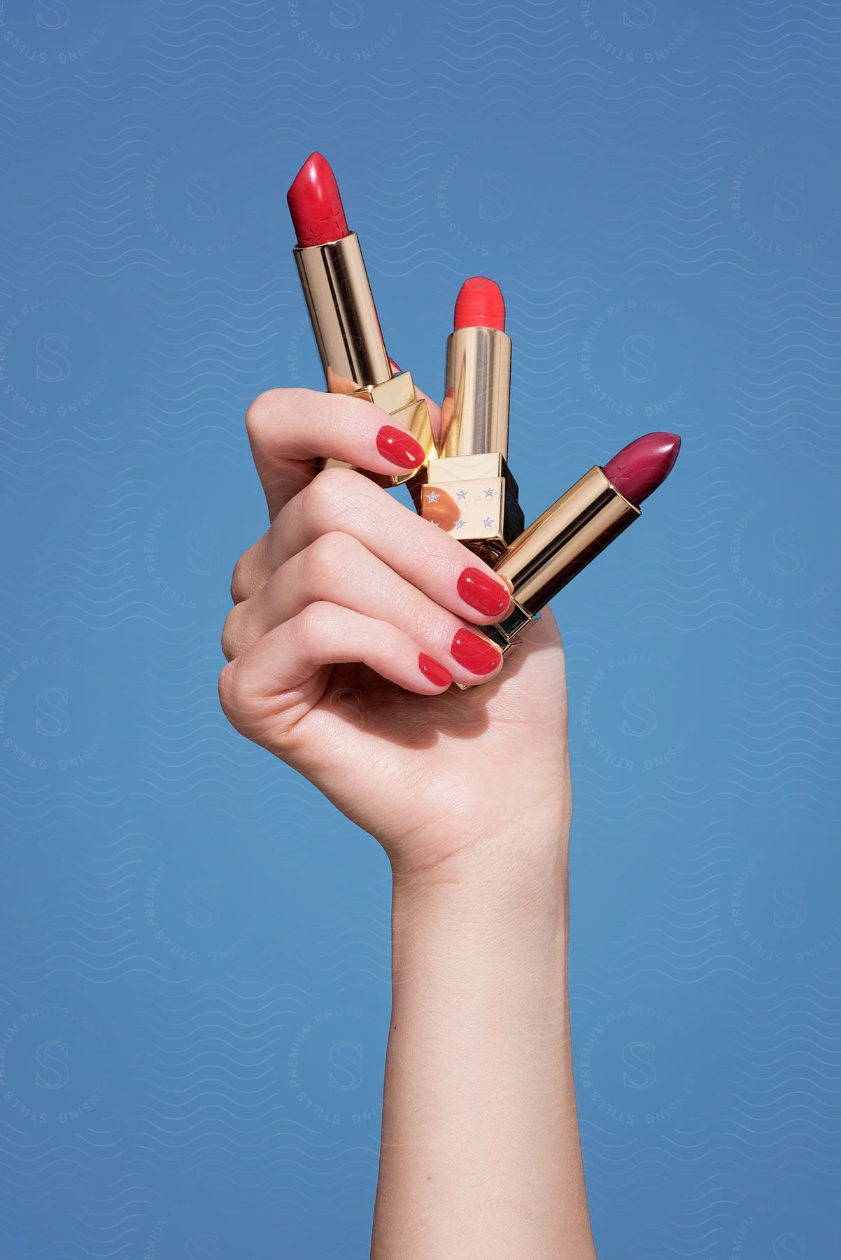 An arm holding three red lipsticks with gold packaging on a completely blue background.