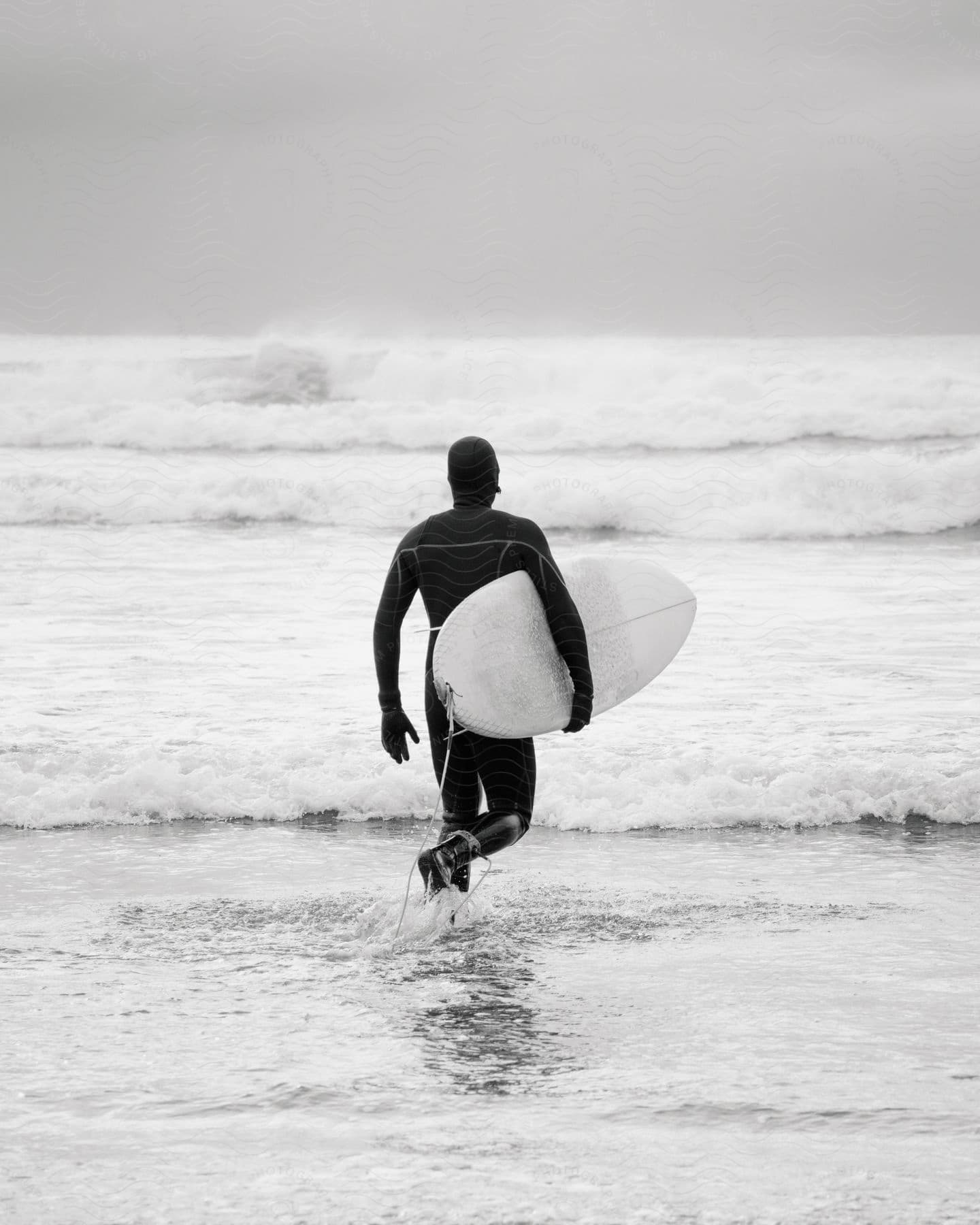 A surfer, board in hand, wades into the ocean as waves crash against the shore.