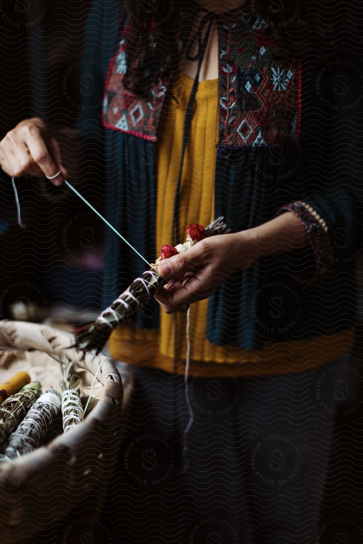 A Native American woman in traditional clothing ties a bundle of sage with rope, surrounded by other bundles of sage in a woven basket.