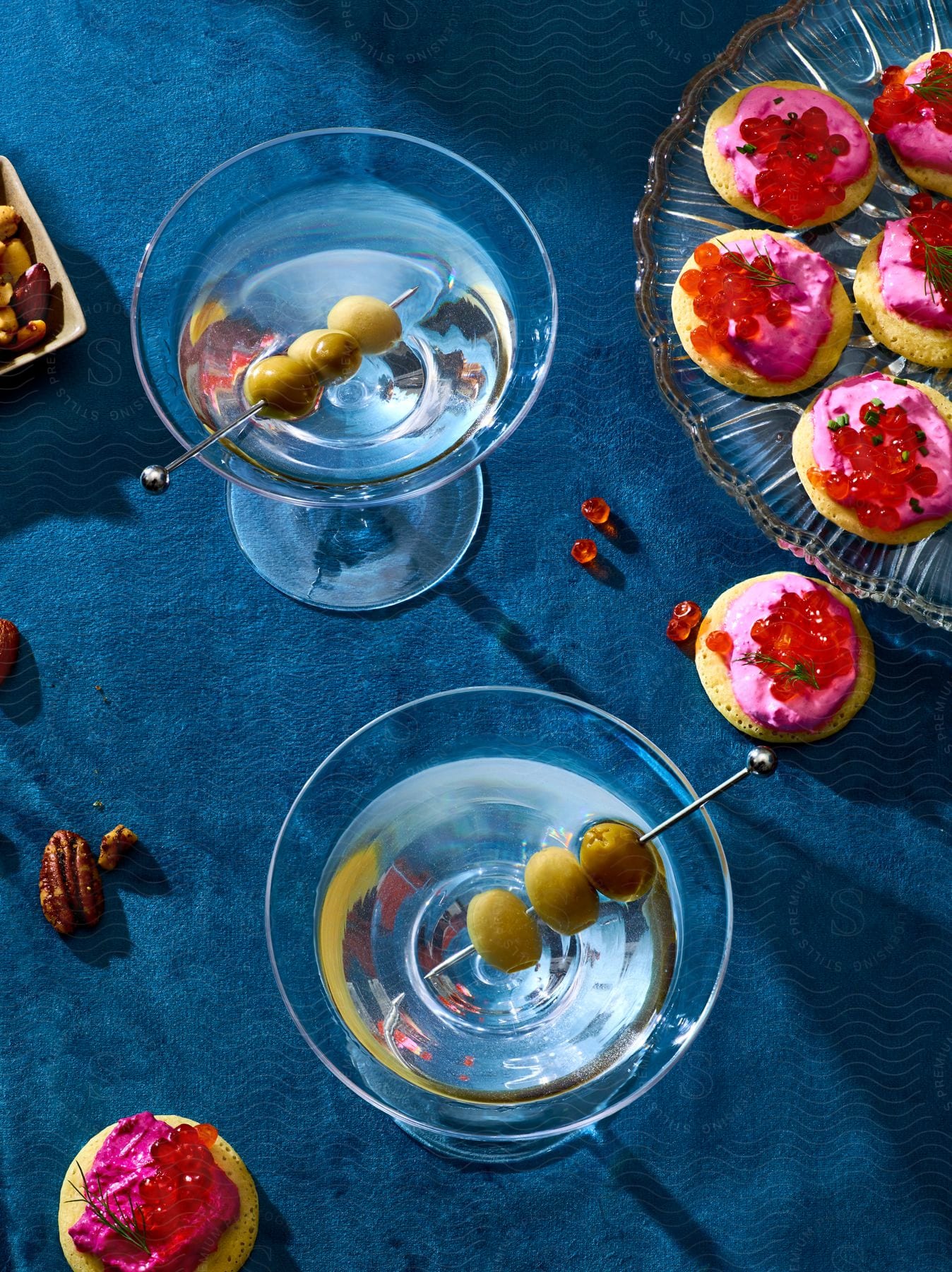 Two bowls with clear liquids and olives inside and a bowl with pink cookies on top.