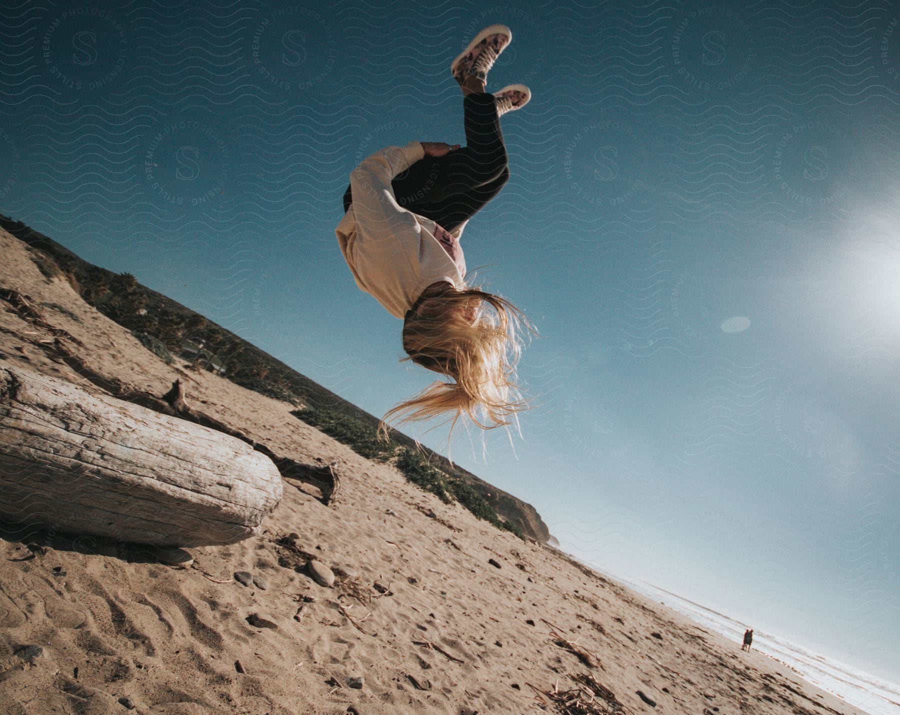 A young blond girl in a long-sleeved shirt flips off a log on the beach on a sunny day