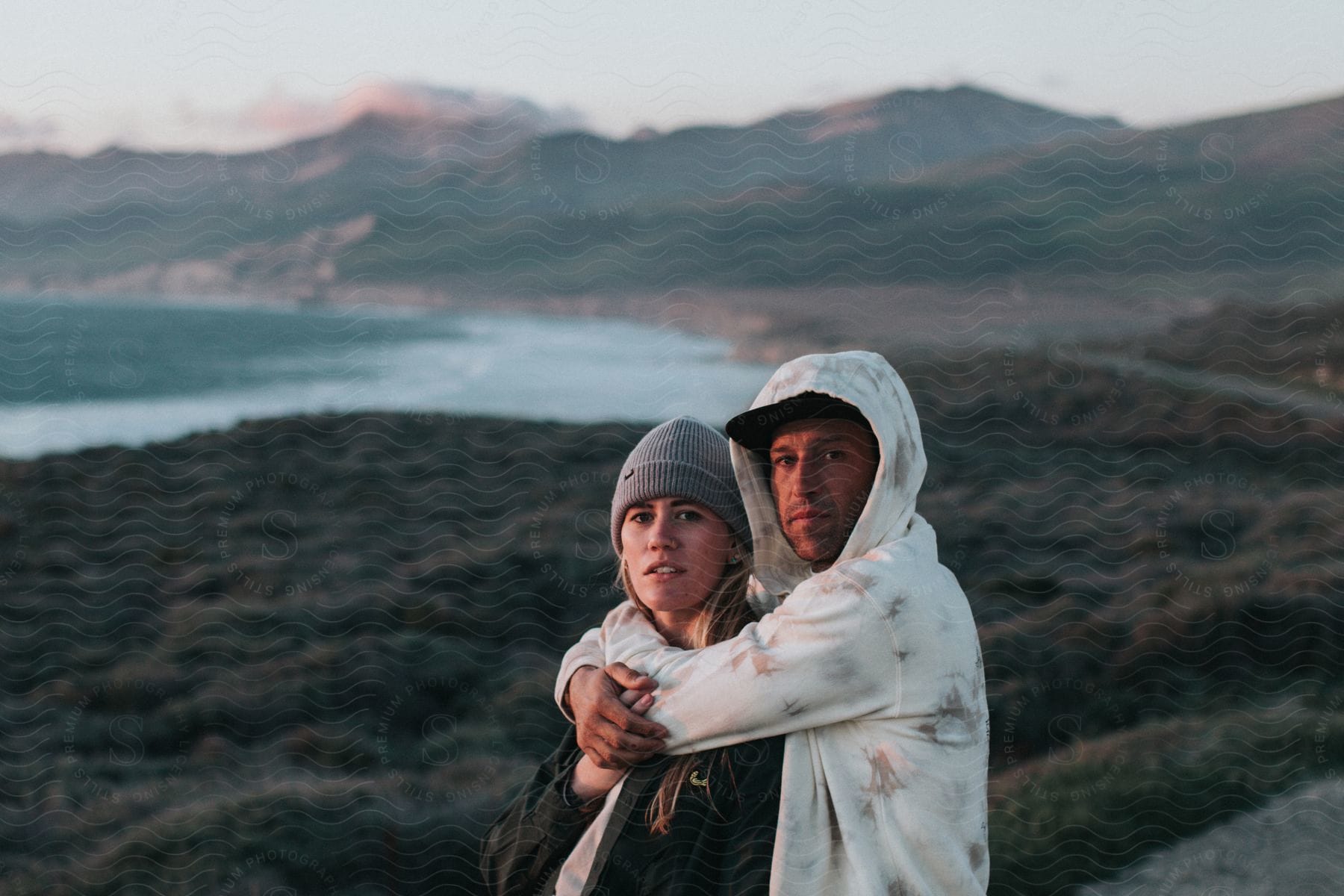 Overlooking the hills and body of water background, a man in a hoodie sweatshirt hugs a lady wearing a beanie cap from behind.