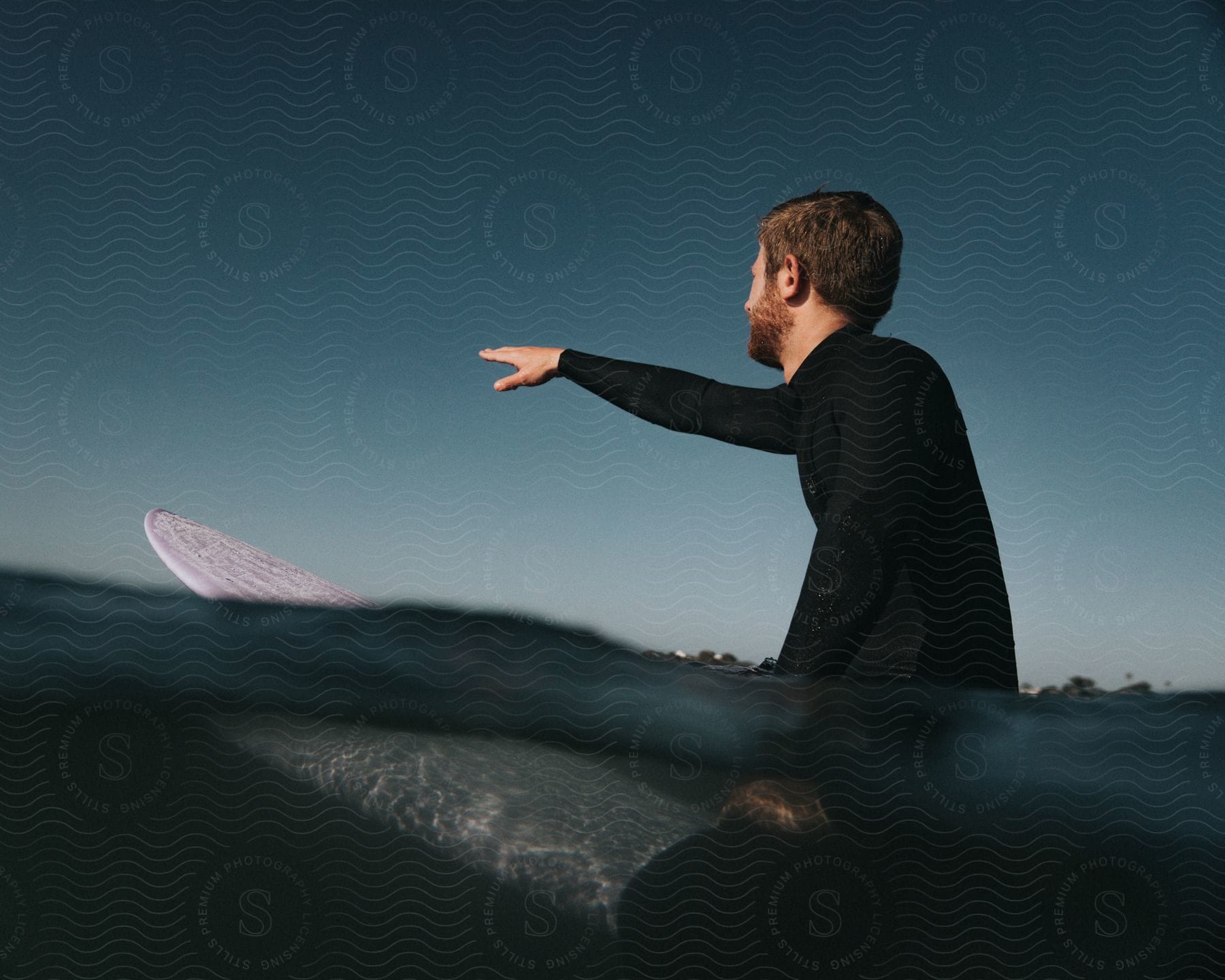 A surfer sitting on a surfboard in a calm sea, pointing out a wave.