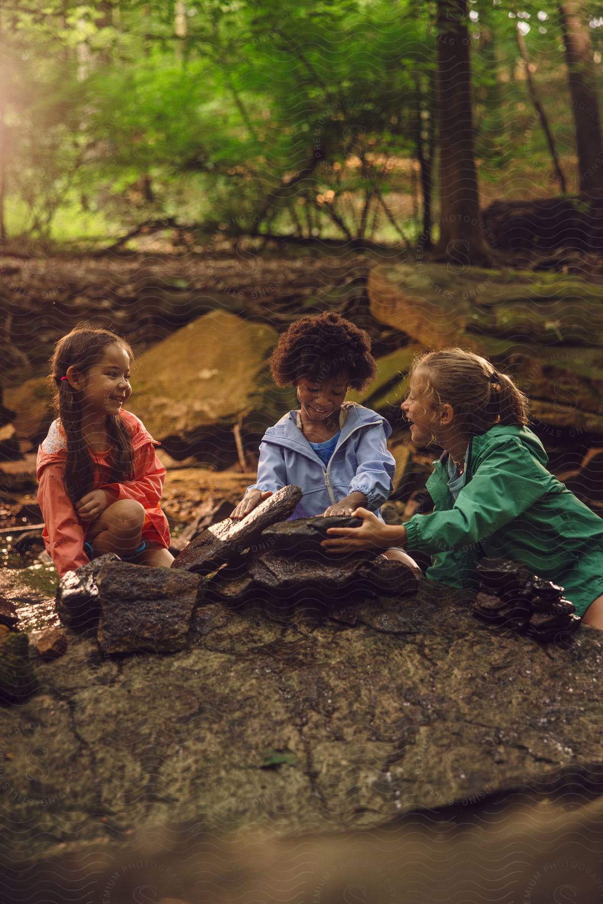 Three children sitting in a forest stacking rocks and laughing with each other.