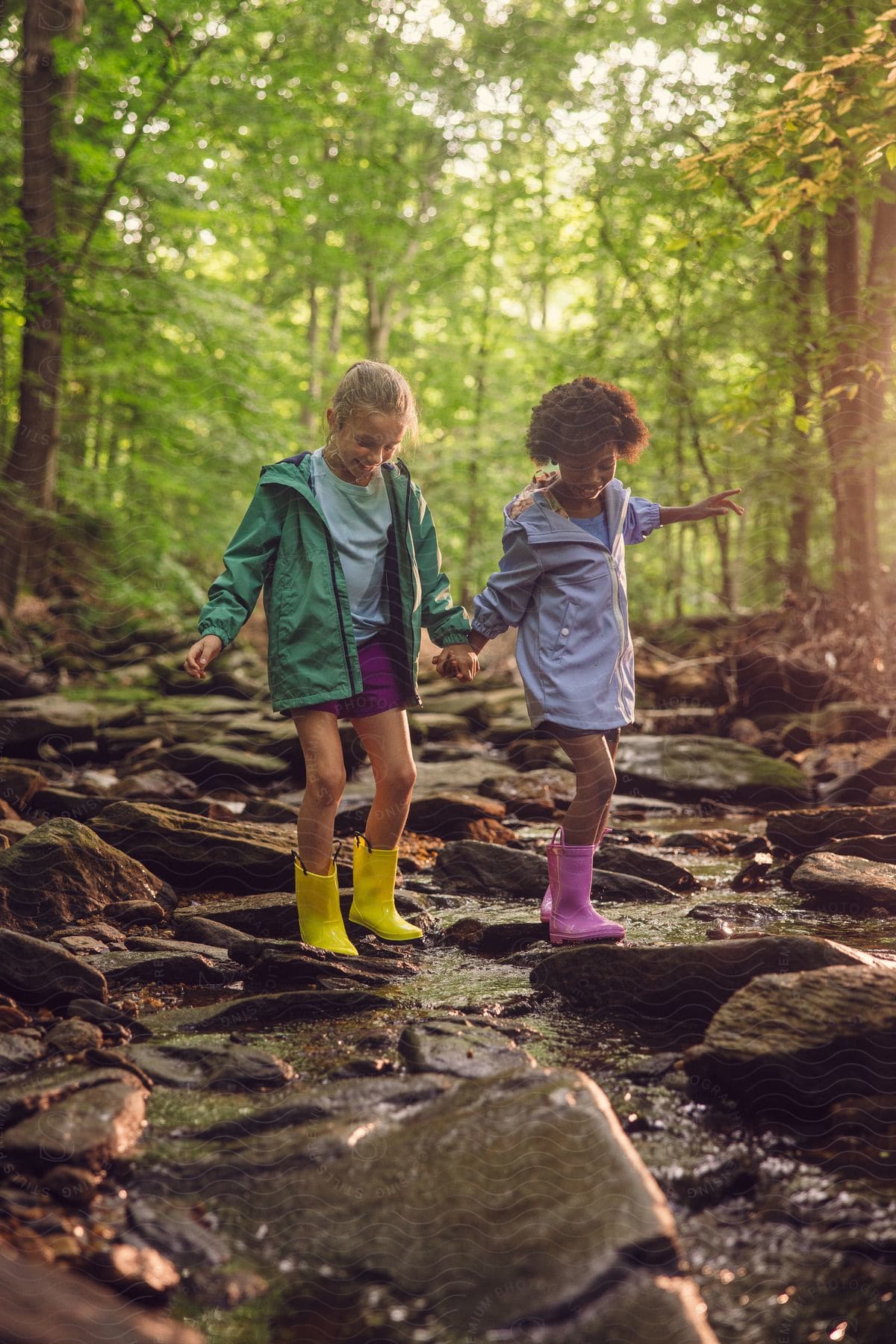 Two young girls are holding hands and walking along a rocky stream in a forest.