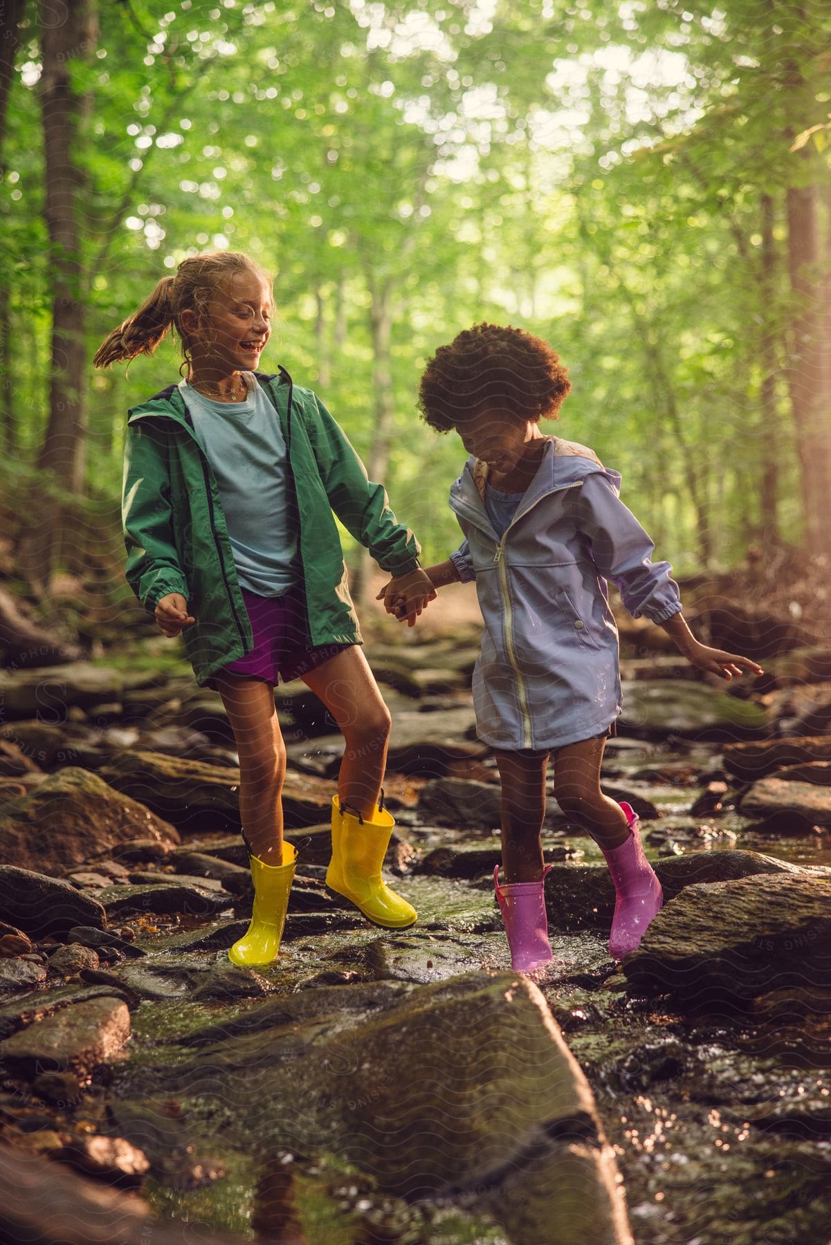 Stock photo of two young girls in rain boots are walking through a stream in the woods together holding hands and smiling