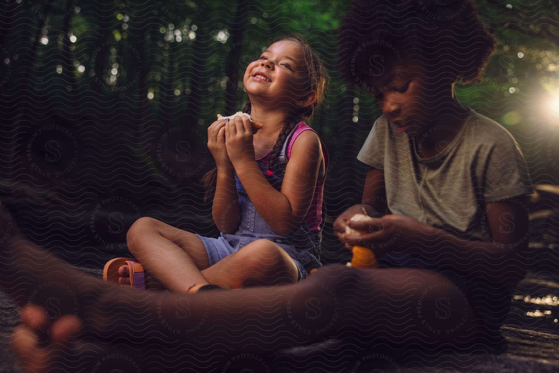 Two little girls are sitting in the woods eating a sandwich as the sun shines through the trees in the distance