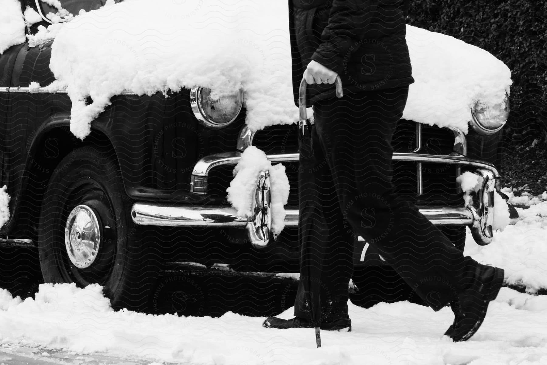 Stock photo of a man uses a cane while walking near an older model automobile covered in snow.