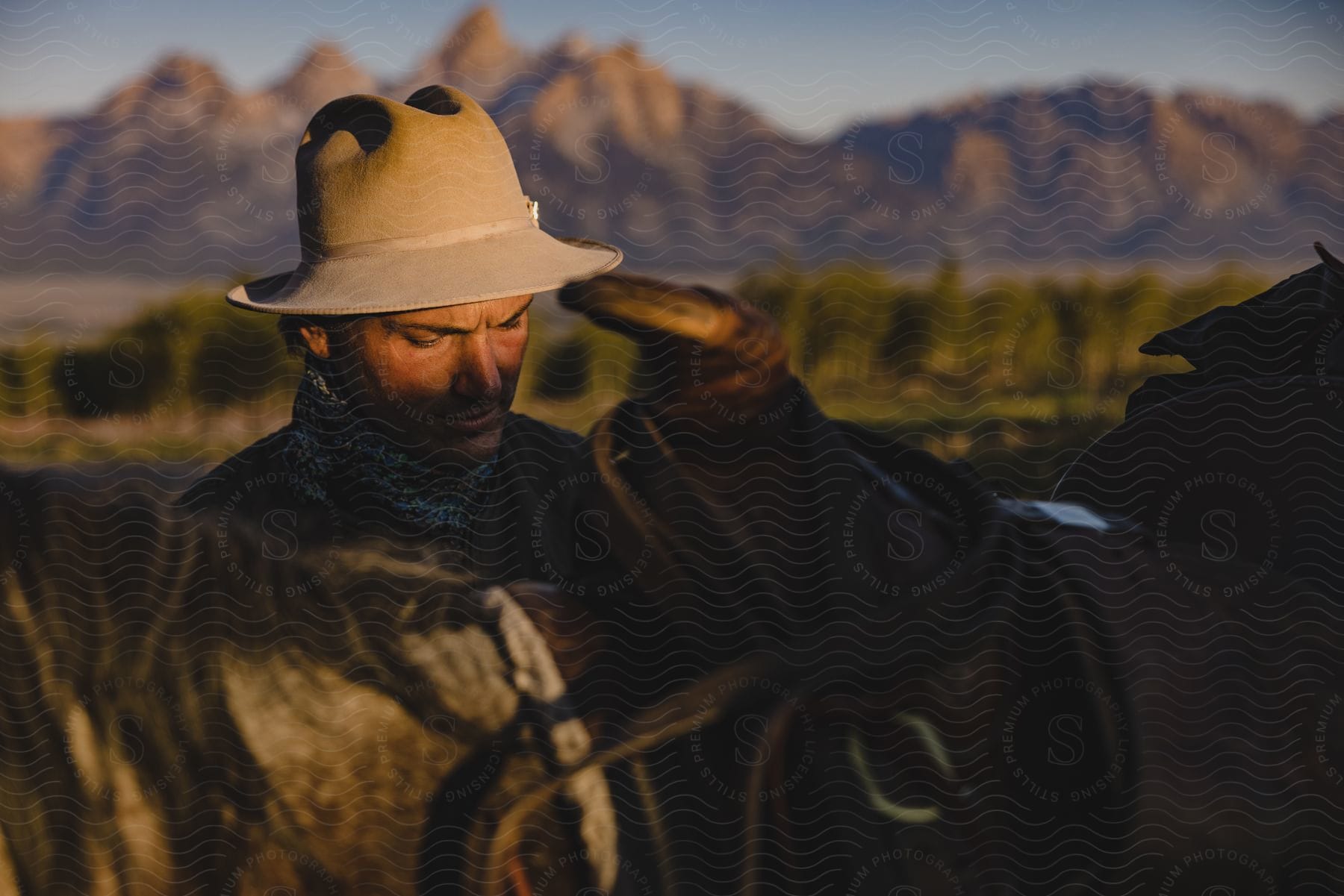 A farmer standing next to his saddled horse as the sun casts shadows across his face with mountains on the landscape behind him