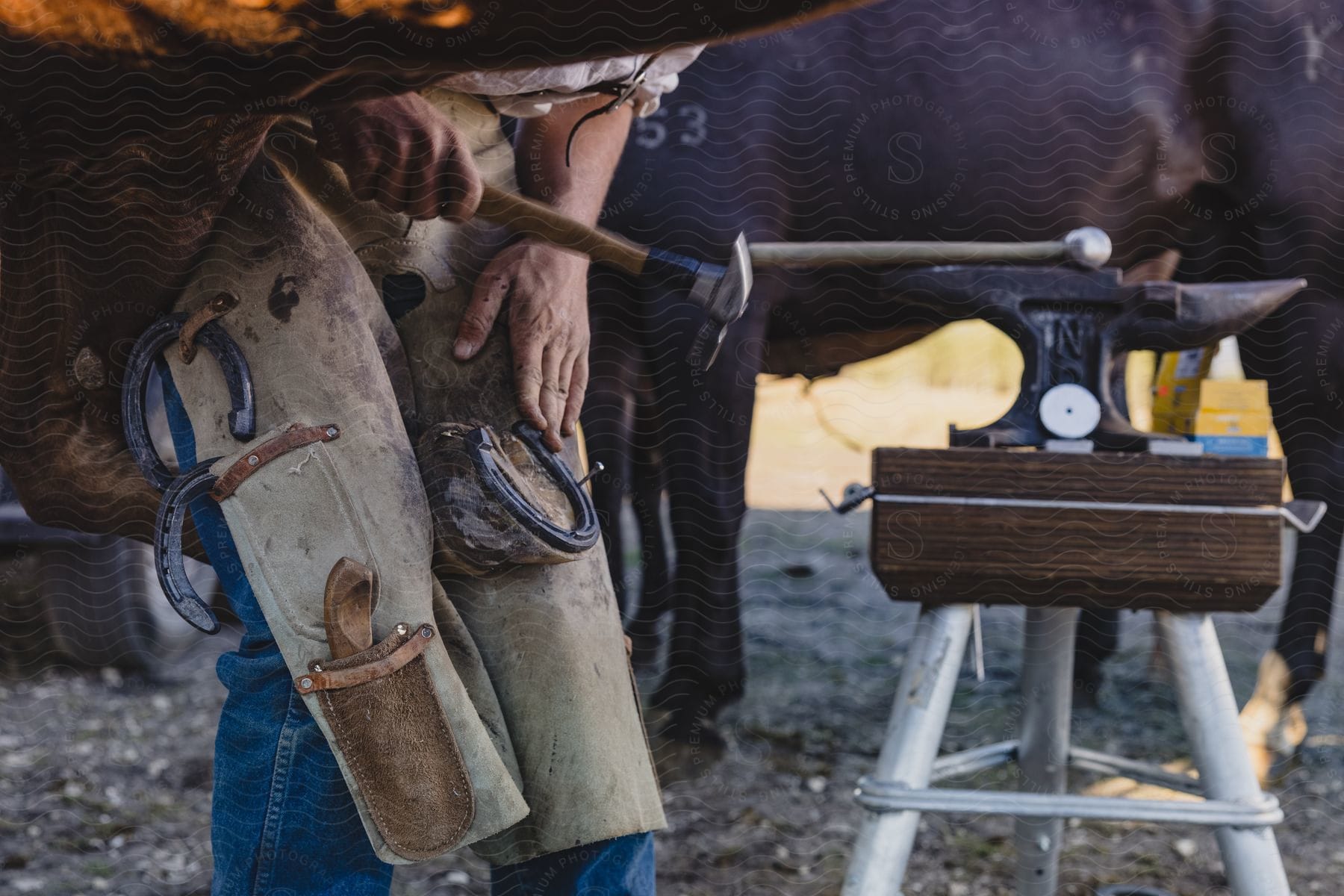 Close up of a man with a horse's hoof between his legs while he hammers a nail into the horseshoe