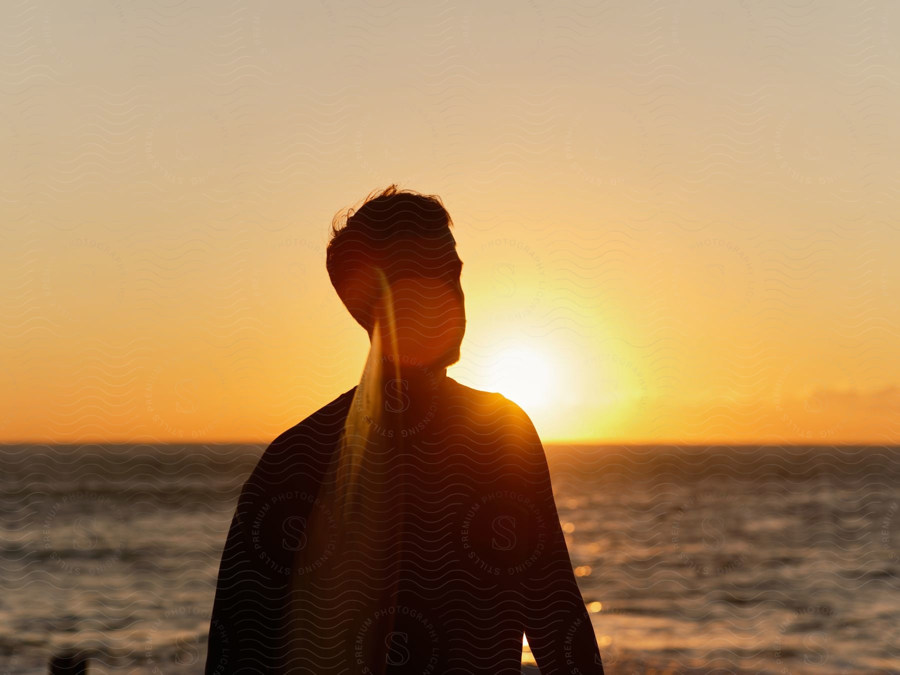 Setting sun back lights man standing near ocean.