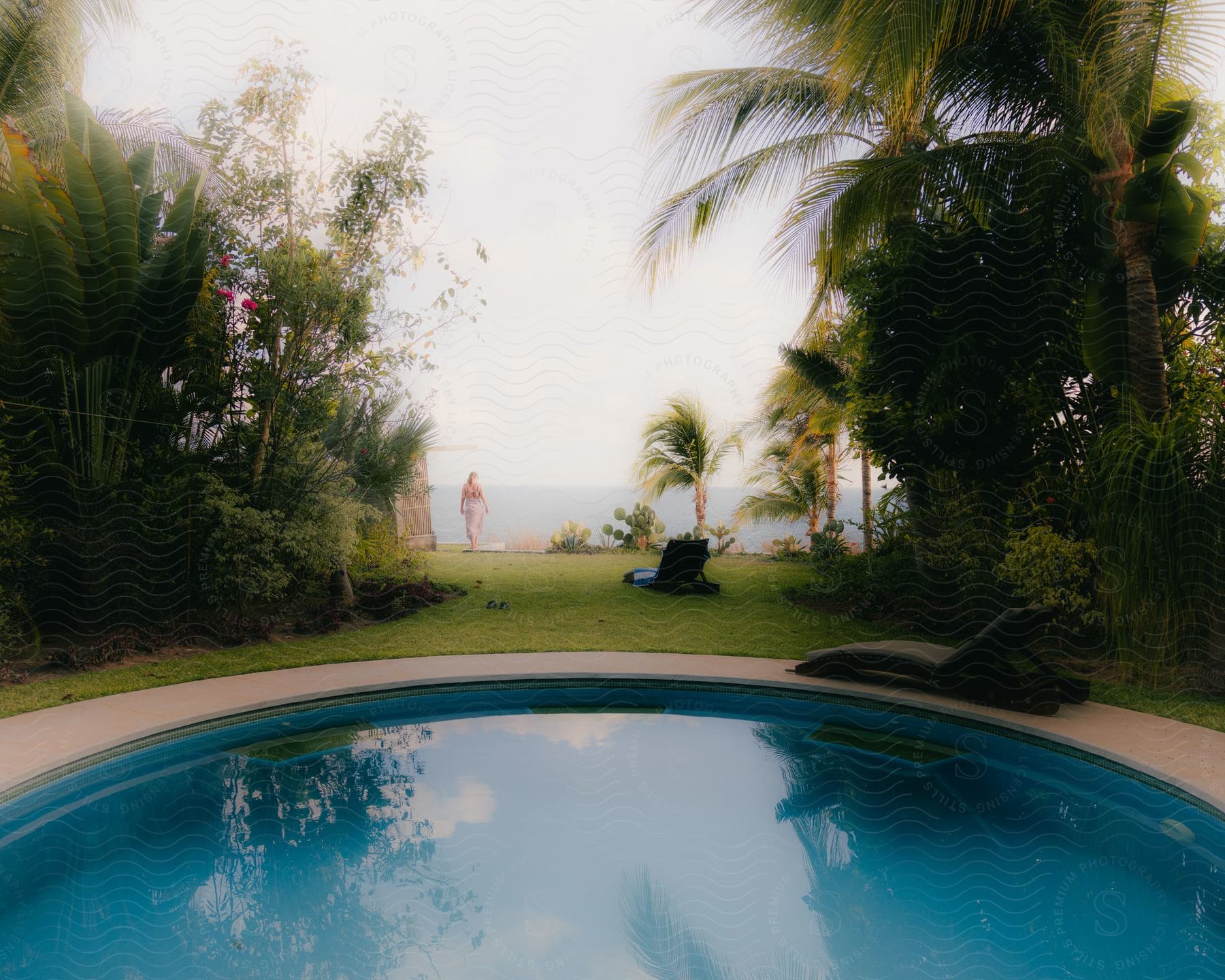 A woman stands further away from swimming pool and trees as she looks at the open ocean.