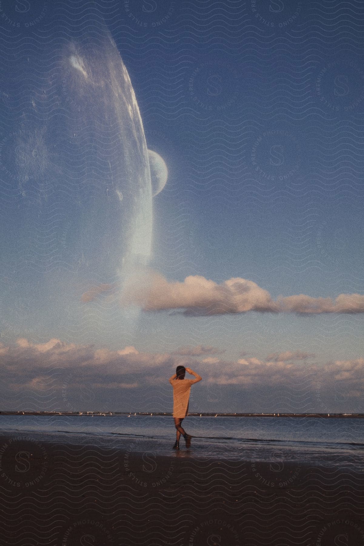 A woman walks along the ocean shore with her hands raised to her forehead under partly cloudy skies.