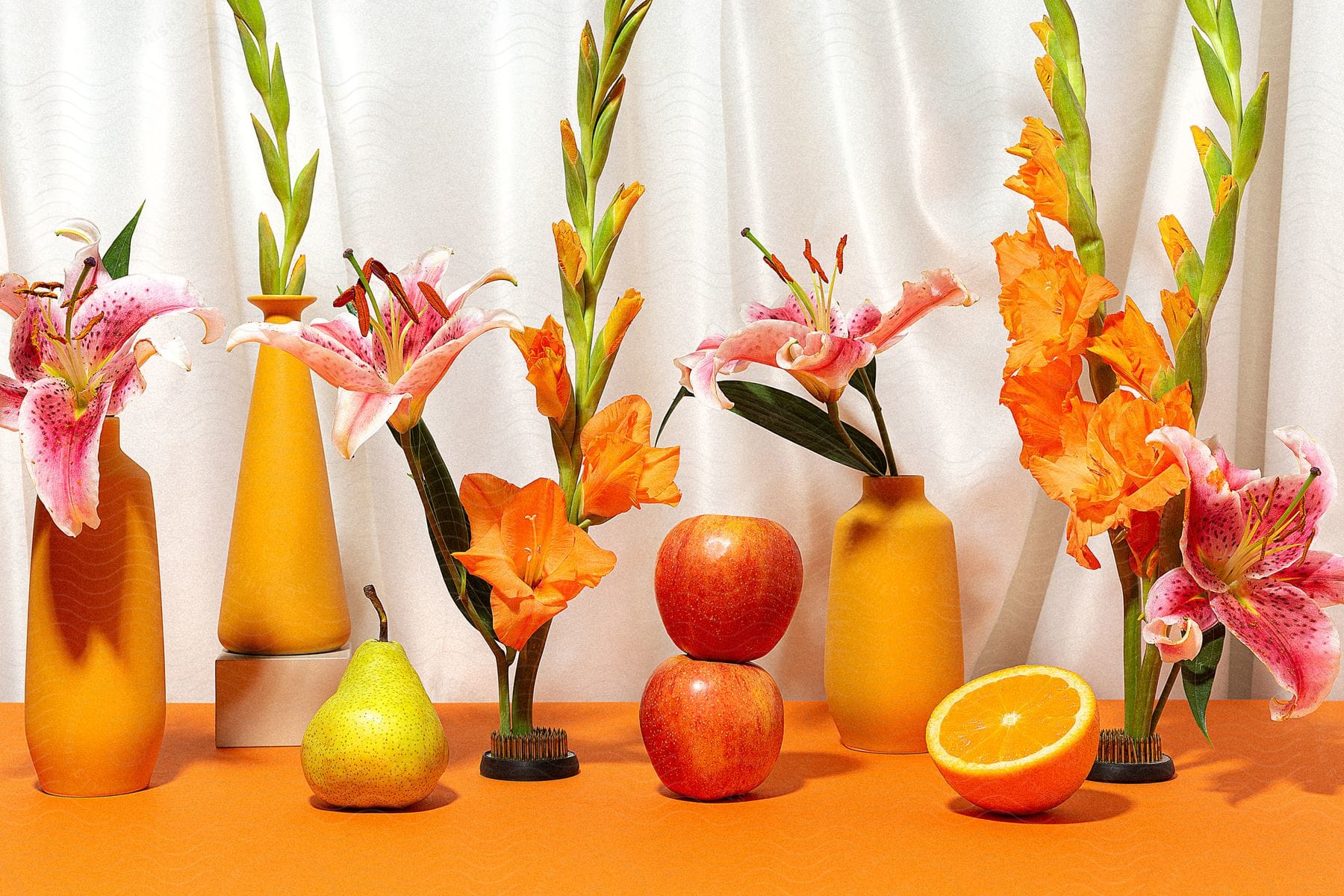 Two apples, an orange and a pear sit on an orange tabletop with vases of lilies.