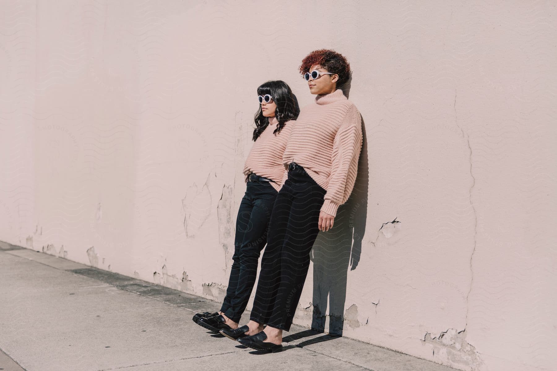 Two women posed leaning on a wall wearing white sunglasses, striped sweaters, black pants and black shoes.