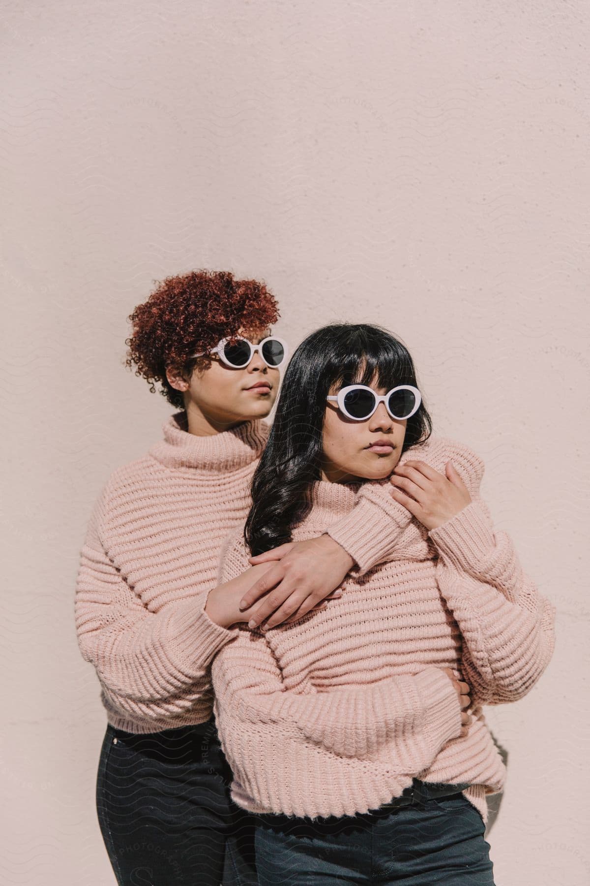 Two women wearing baggy pink sweatshirts and sunglasses are standing together and one woman has her arms around the other