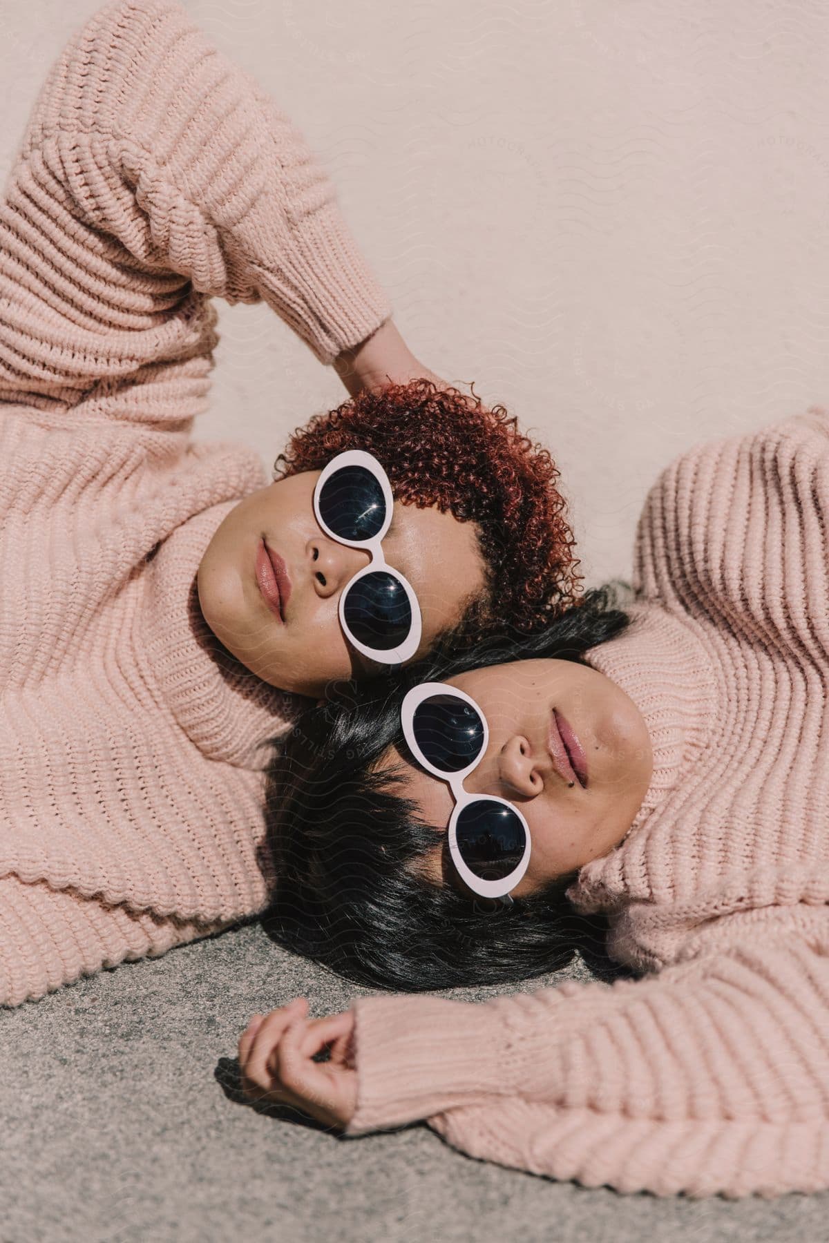 Two women wearing baggy pink sweatshirts and sunglasses are lying on the floor with their heads together from opposite directions