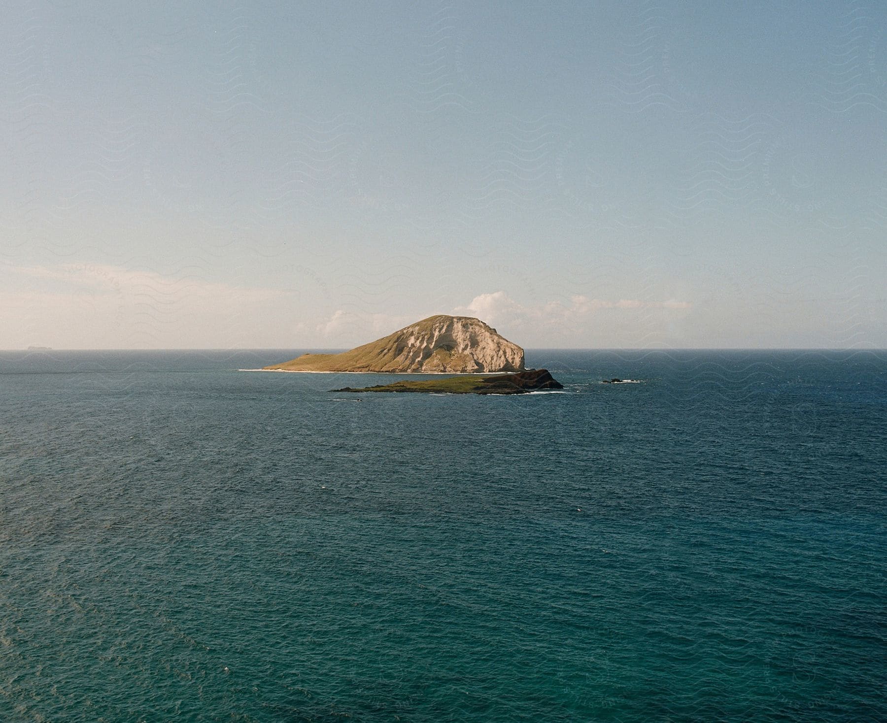 A large island of vegetation and rocks isolated in the middle of the blue ocean on a clear day.