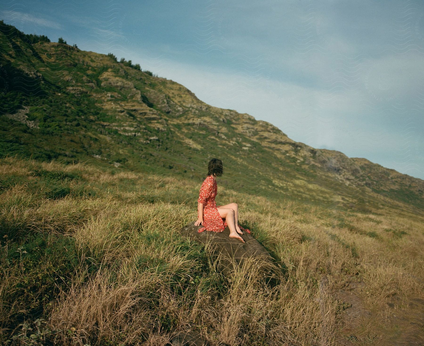 A barefoot woman in an orange dress sits on a rock in the grasslands below a mountain ridge.