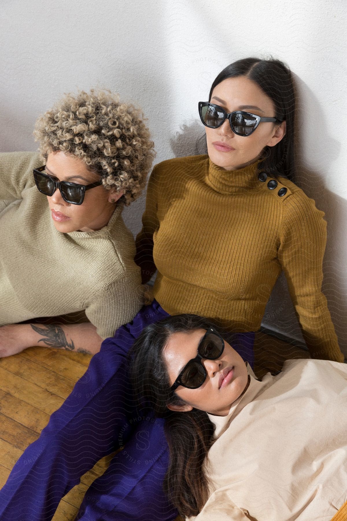 Three women leaning against each other on the floor, wearing fashionable sunglasses.