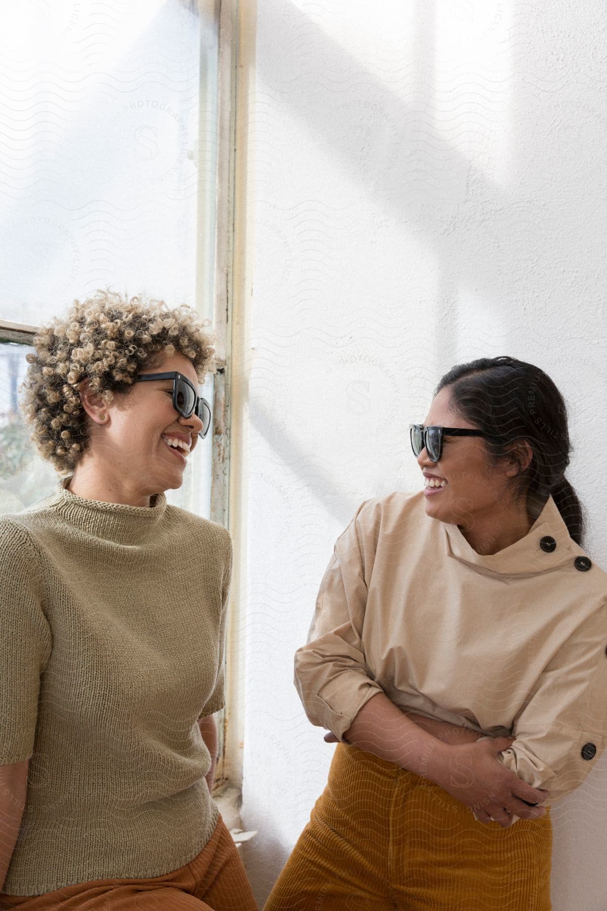 Woman with curly hair sunglasses and brown sweater and pants laughs with woman with sunglasses beige shirt and brown pants