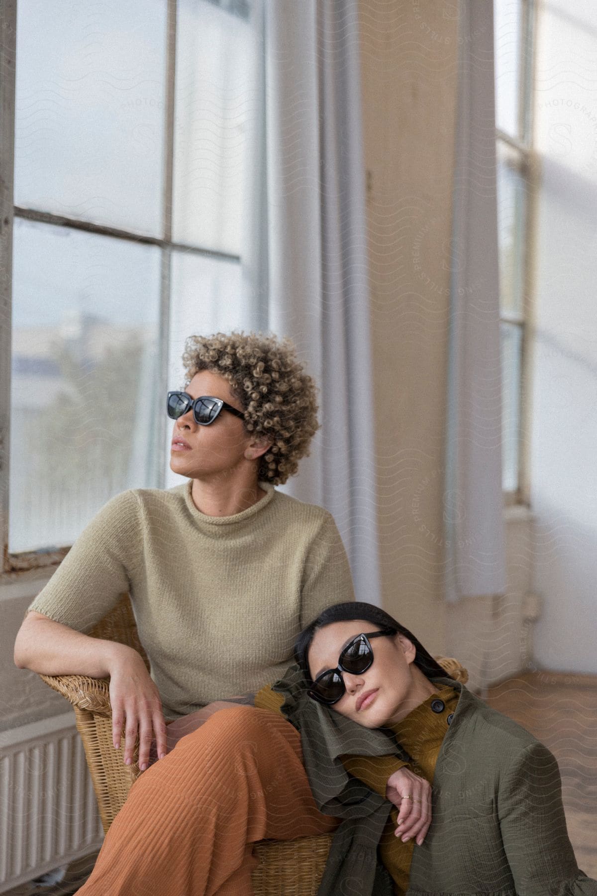 Woman with dark hair sunglasses and blazer jacket sits by woman's legs wearing brown pants short sleeve sweater and sunglasses sitting in armchair