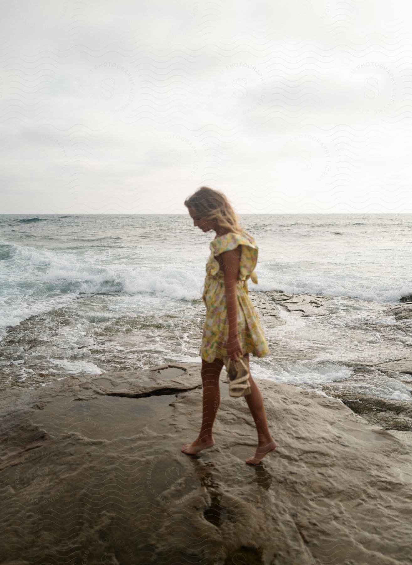 A blonde woman in a yellow sun dress walks barefoot on a rocky beach.