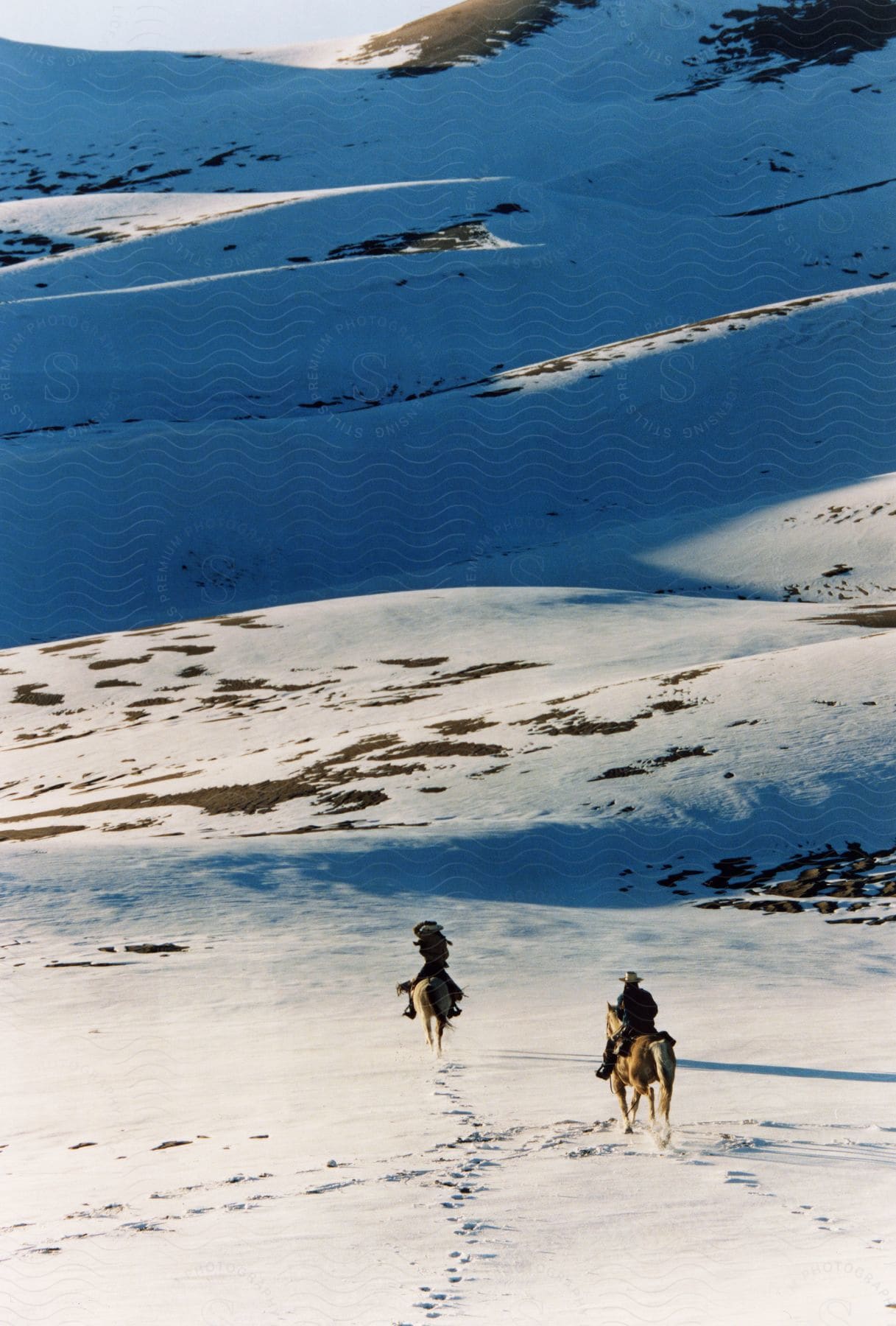 Two people riding horses in a mountain ranch land area covered in snow.