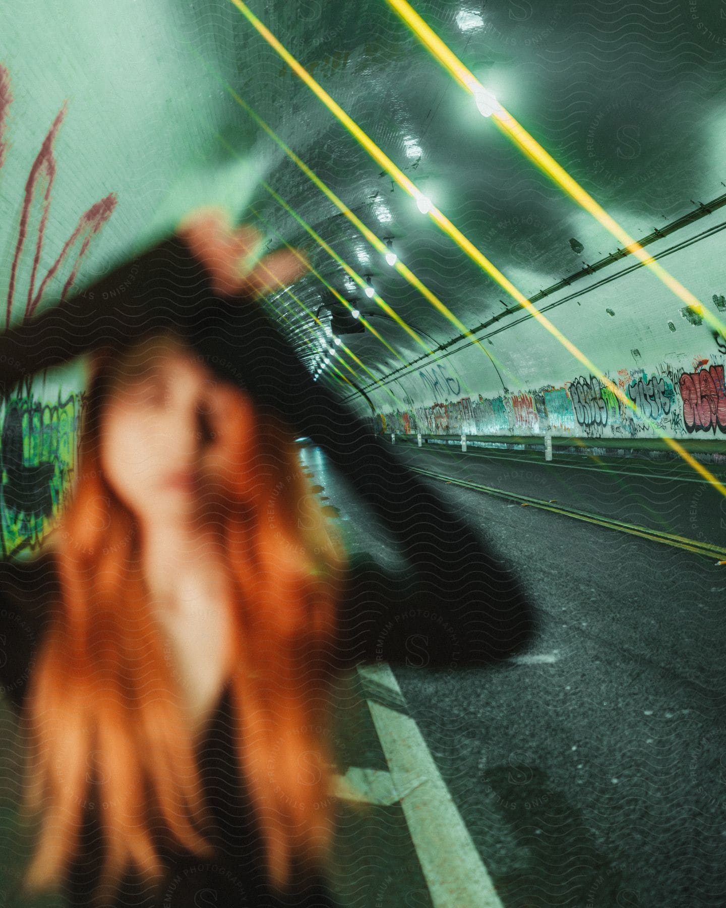 An out-of-focus woman with red hair and long sleeves stands in a graffiti-covered tunnel, illuminated by the light of street lamps.