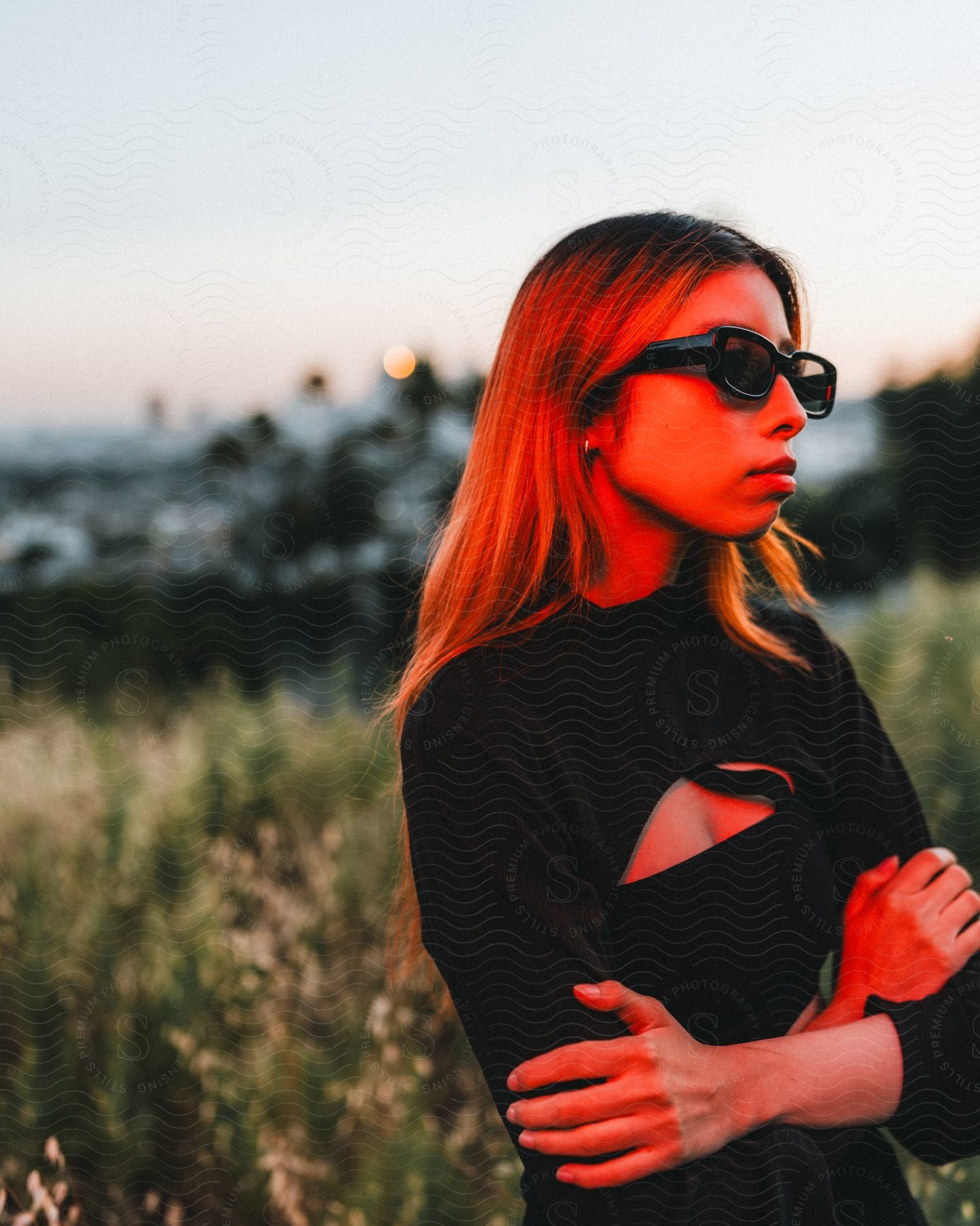 Woman with brown hair sunglasses and black shirt stands with arms crossed in red lighting in the field