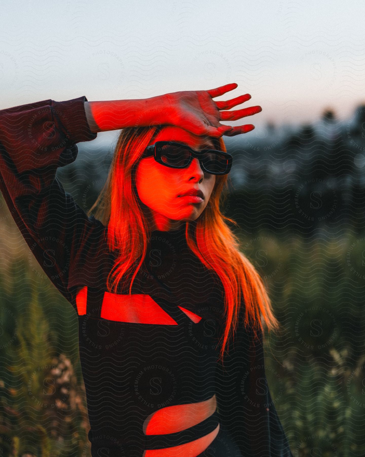 Red light shines on a teenage girl wearing sunglasses and a black outfit with her hand on her forehead as she stands outside